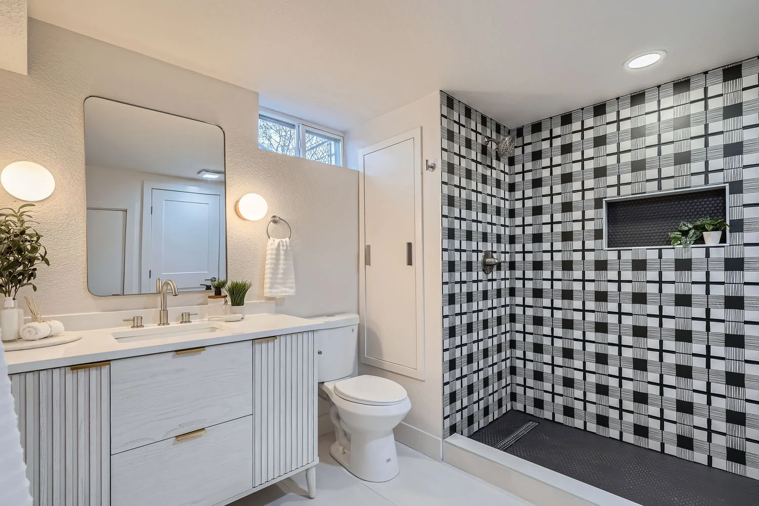Modern bathroom featuring a white vanity with a large mirror, a toilet, and a walk-in shower with black and white geometric tiles and built-in shelving for decor.
