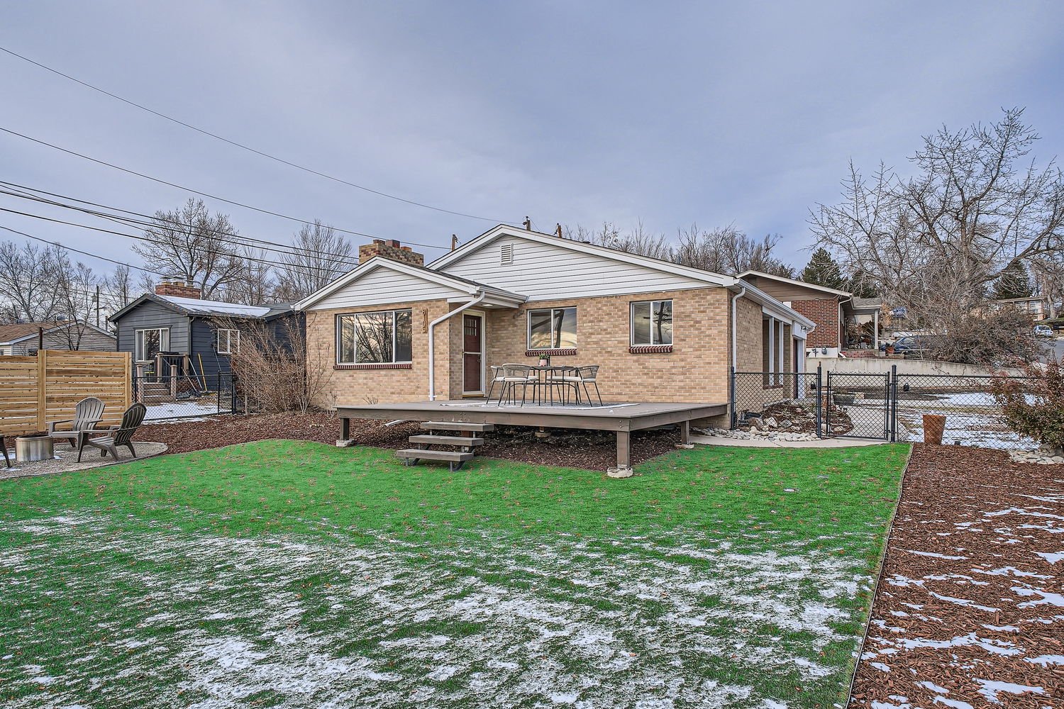 Backyard with a green lawn patch and a raised wooden deck with outdoor furniture, snow on the ground, a chain-link fence, and neighboring houses under a cloudy sky.