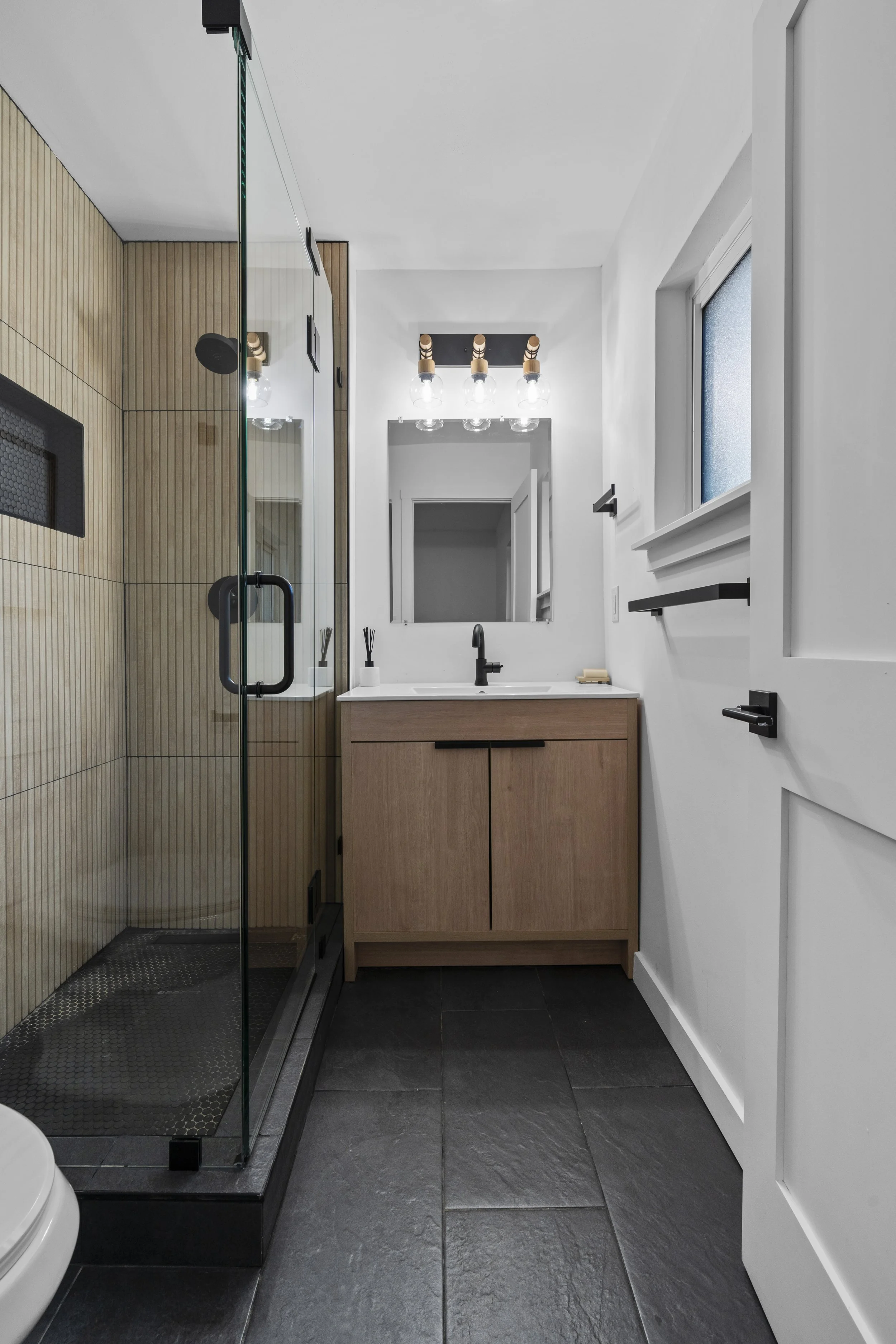 Modern bathroom with a glass shower enclosure, wooden vanity with a white countertop, black faucet, large mirror, black light fixture, frosted window, black door handle, black towel holder, and dark slate tile flooring.