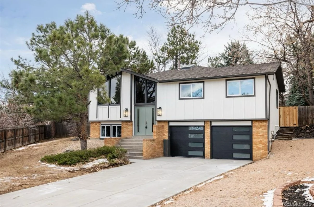 A modern two-story house with a front yard, featuring a brick and white exterior, large front windows, and a two-car garage with black doors, in a suburban neighborhood.