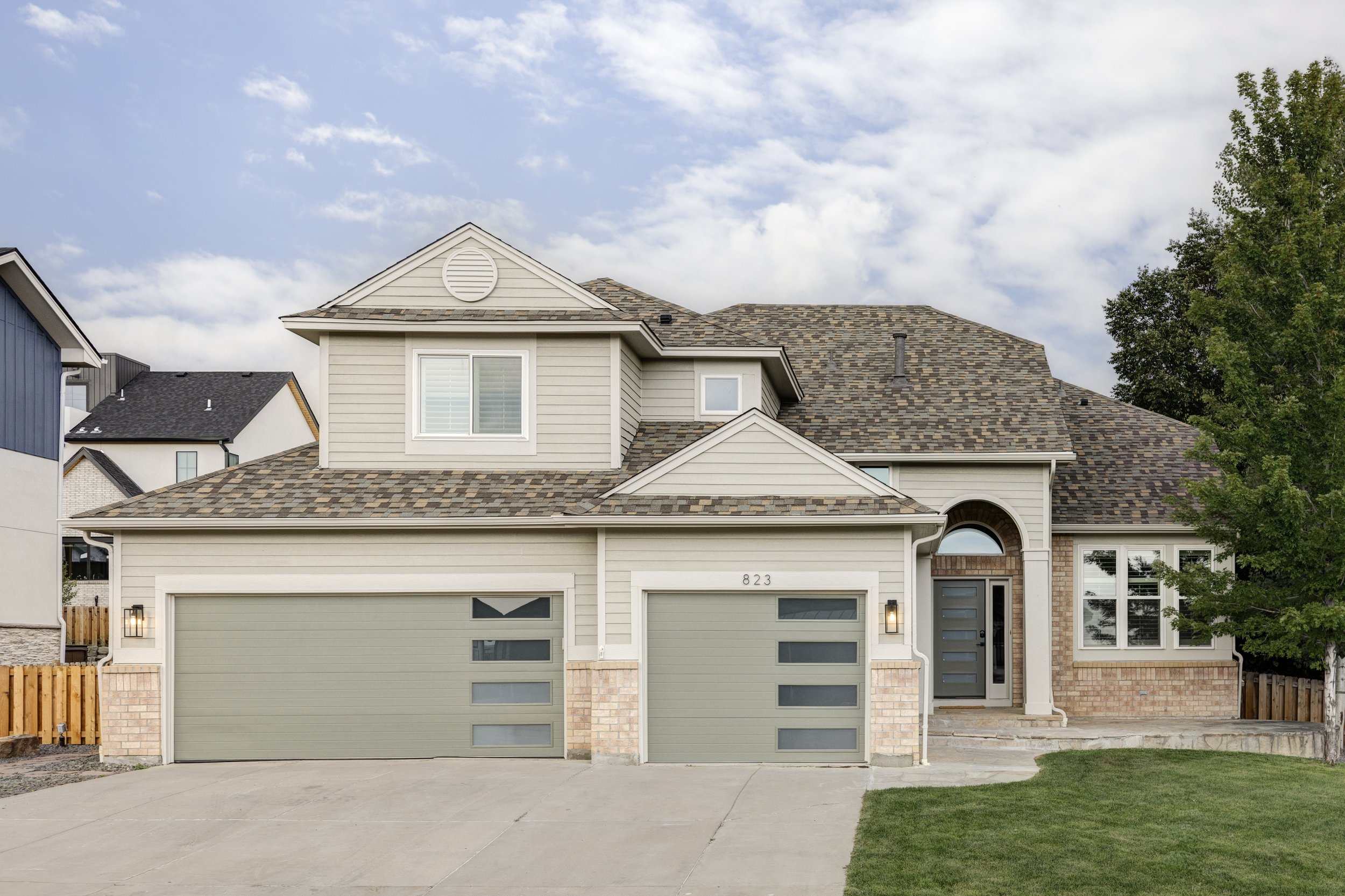 Front view of a modern two-story house with a double garage, brick and siding exterior, paved driveway, and a green lawn, under a partly cloudy sky.