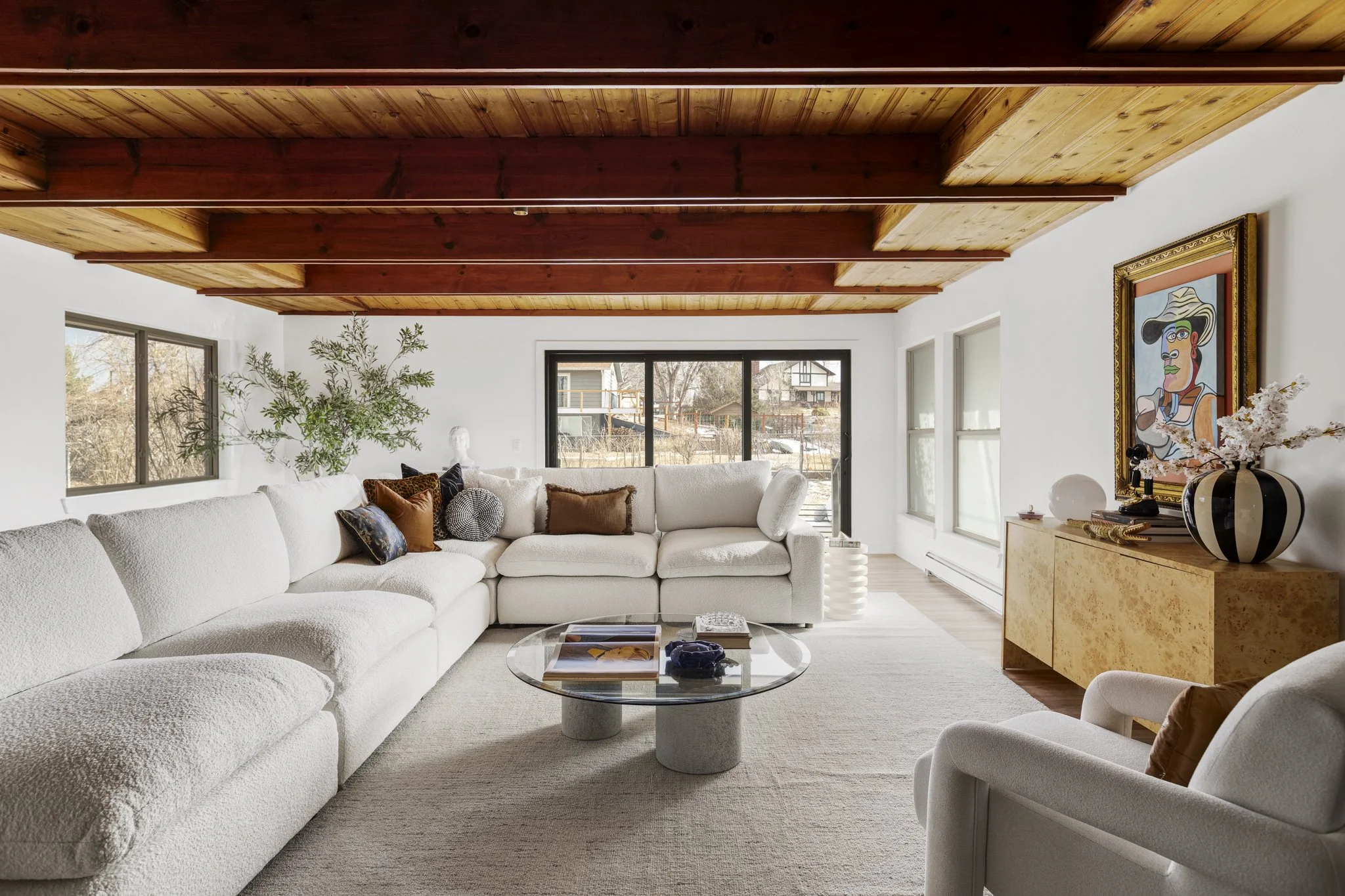 Living room with white sectional sofa, armchair, glass coffee table, artwork on wall, and wooden ceiling in a mid-mod home.