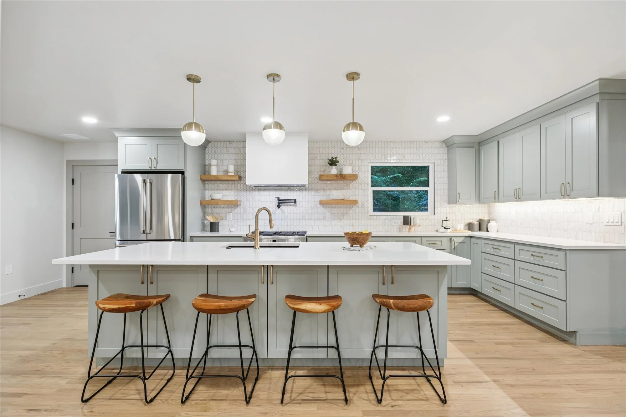 Modern kitchen with light gray cabinets, white countertops, and wooden bar stools at a kitchen island. Includes stainless steel appliances, floating shelves, a small window, and decorative items, with hardwood flooring.