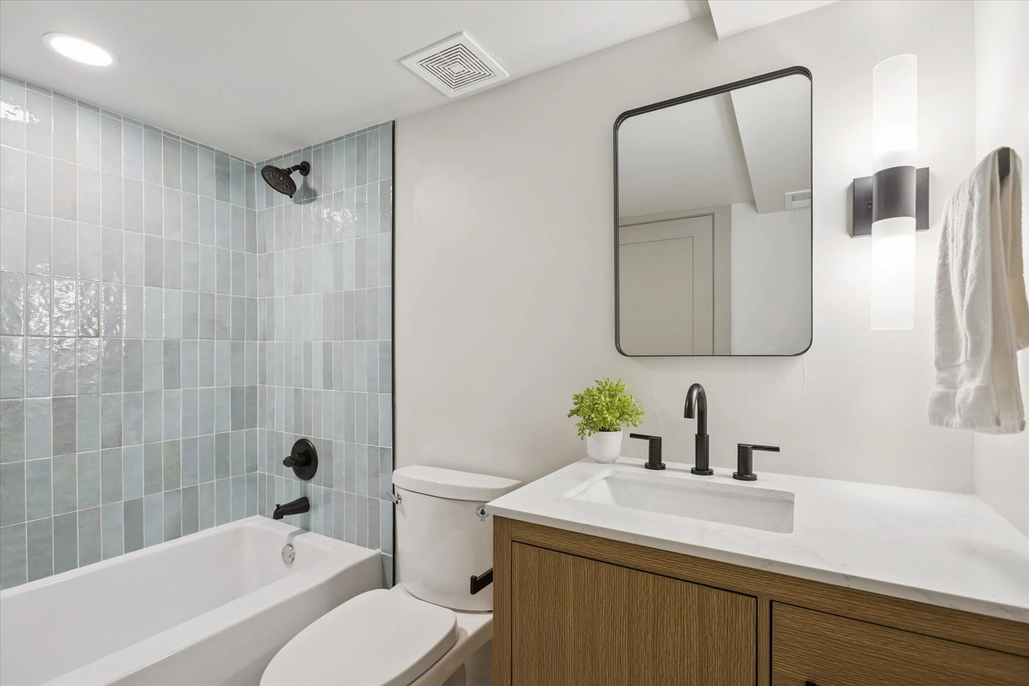 Modern bathroom featuring a walk-in shower with black fixtures, a white bathtub, a toilet, a wooden vanity with a white marble countertop, a black faucet, a small potted green plant, a rectangular mirror, and a wall-mounted light fixture.