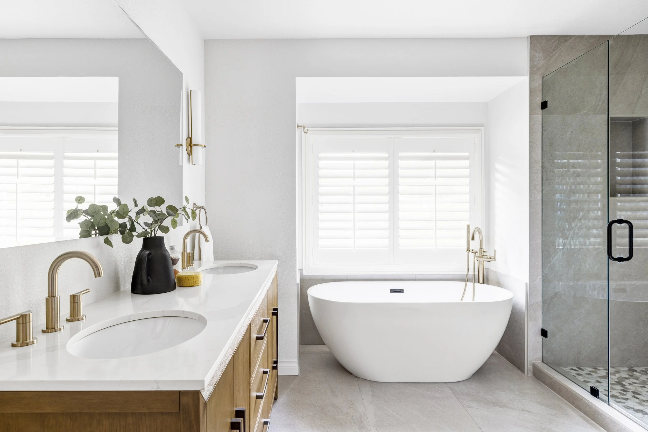 Modern bathroom with double vanity with gold fixtures, black vase with greenery, a white bathtub below a window with white shutters, and a glass walk-in shower with beige tiles.