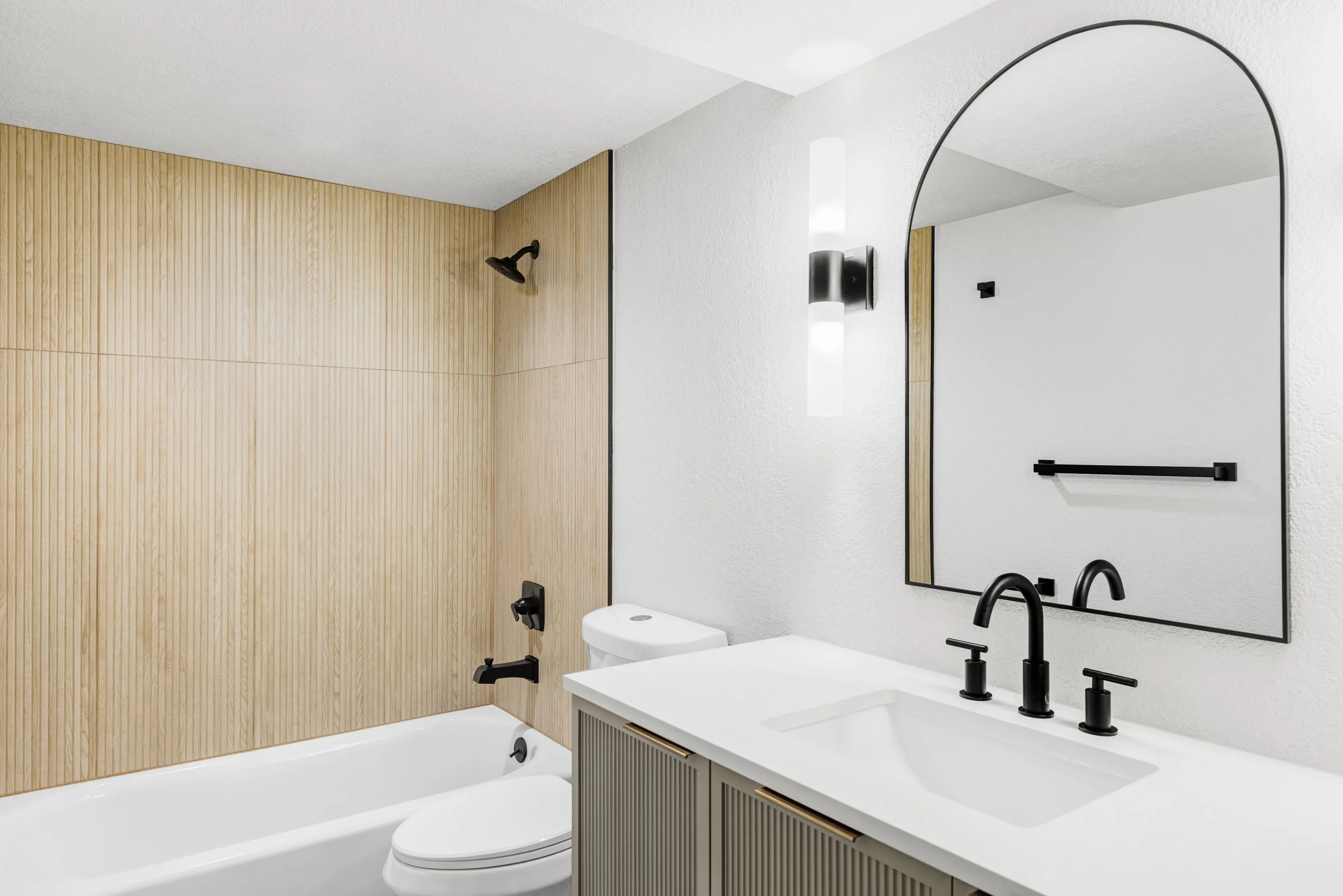 Modern bathroom with a white bathtub, black fixtures, a toilet, a large mirror, a white vanity with black faucet, and a wall-mounted black light fixture.