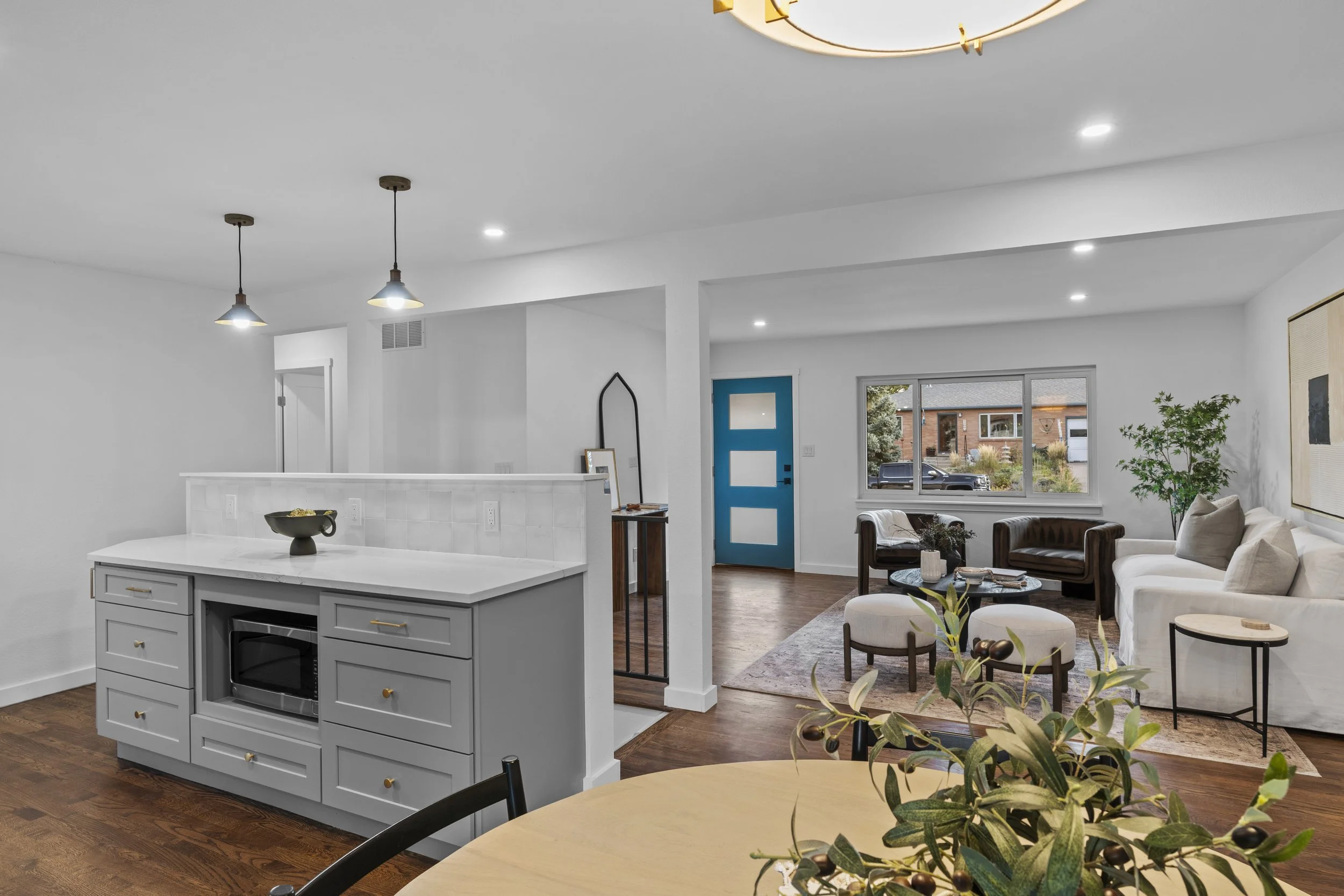 Open-concept living room and kitchen with white walls, hardwood floors, modern furniture, and a blue front door, with a large window showing a street view.