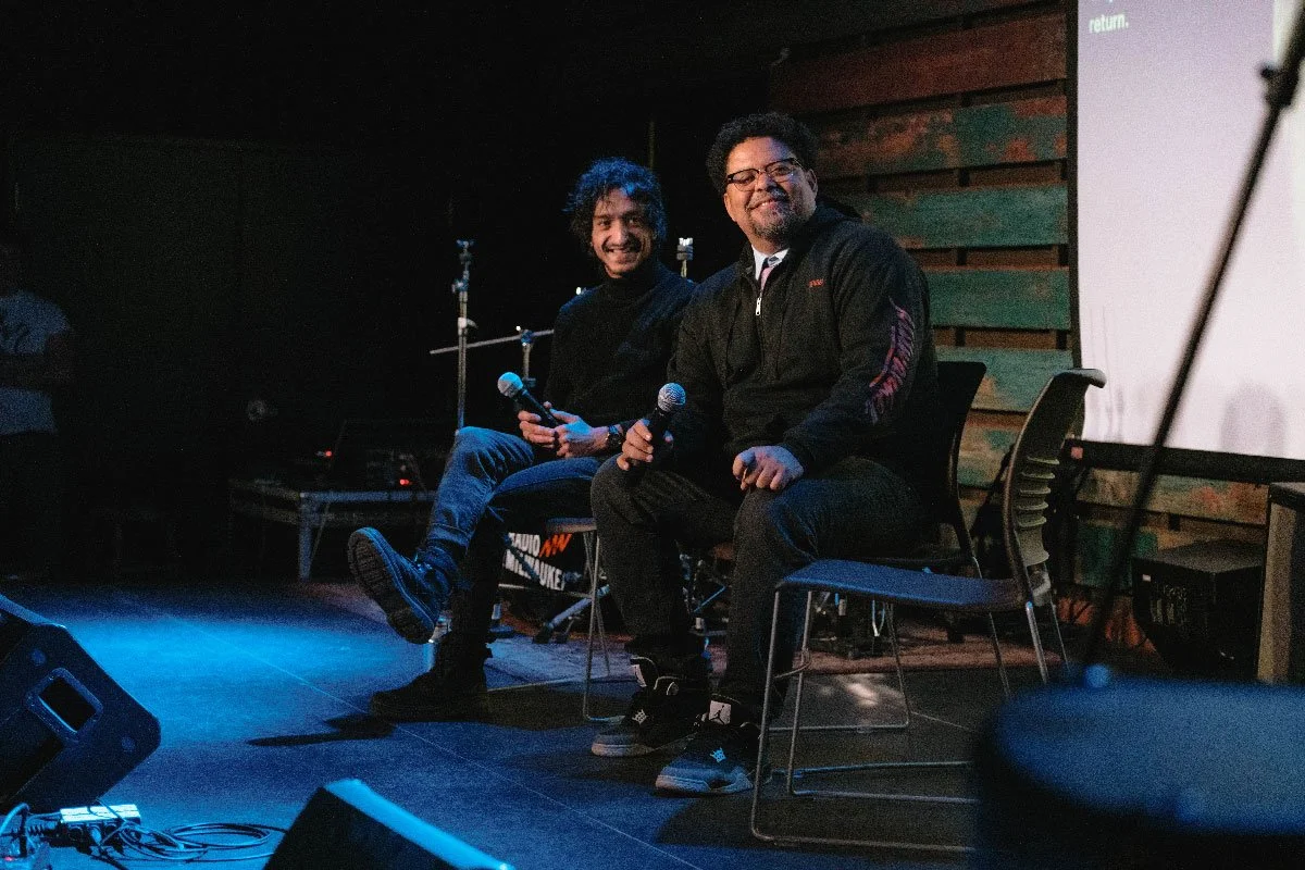 Two men sitting on stage, holding microphones, smiling, with a wooden panel wall and a large screen behind them.