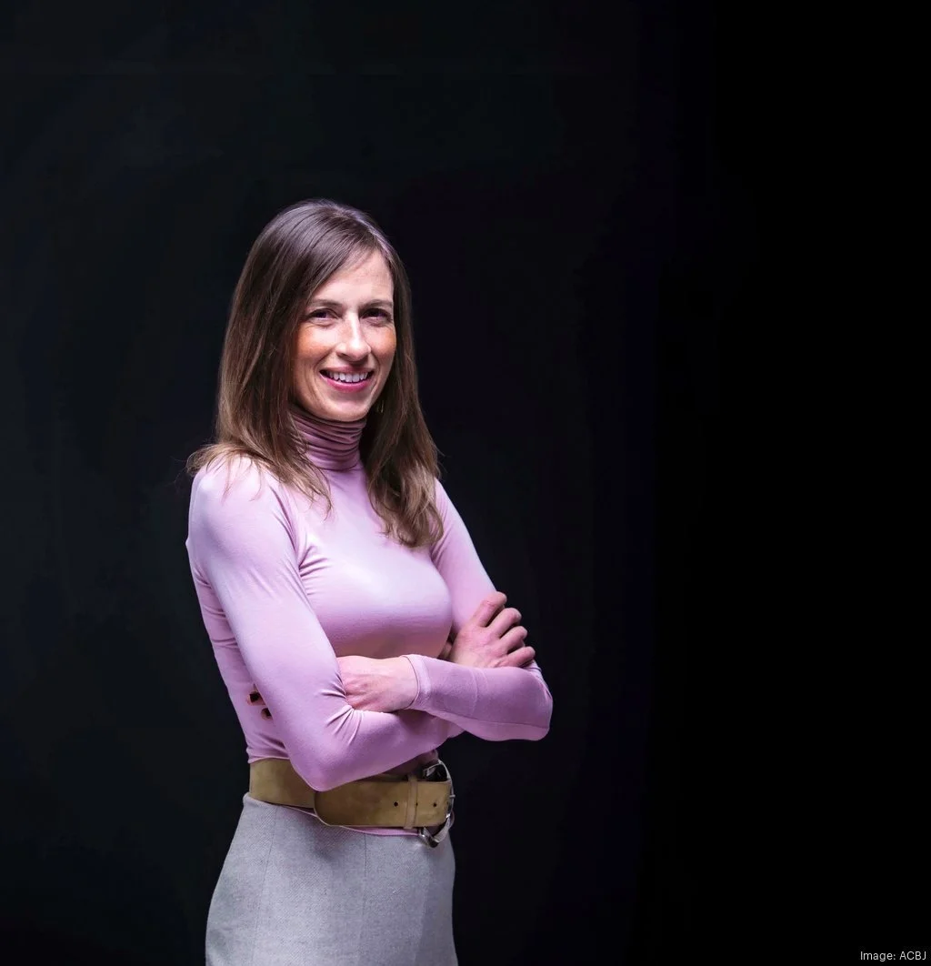 Portrait of a woman with shoulder-length brown hair, wearing a pink long-sleeve turtleneck and beige pants, standing with arms crossed in front of a dark background.