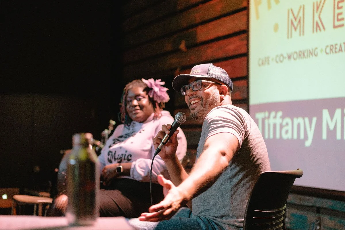 Two people sitting on a stage, one man holding a microphone and smiling, and one woman with a flower in her hair, listening. A screen behind them displays the name Tiffany M.