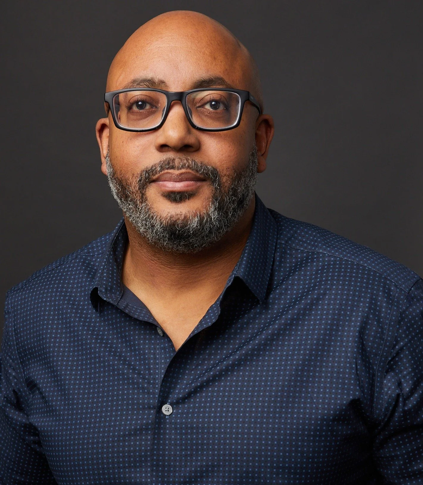 A portrait of a man with glasses and a beard, wearing a dark blue patterned shirt, against a dark background.