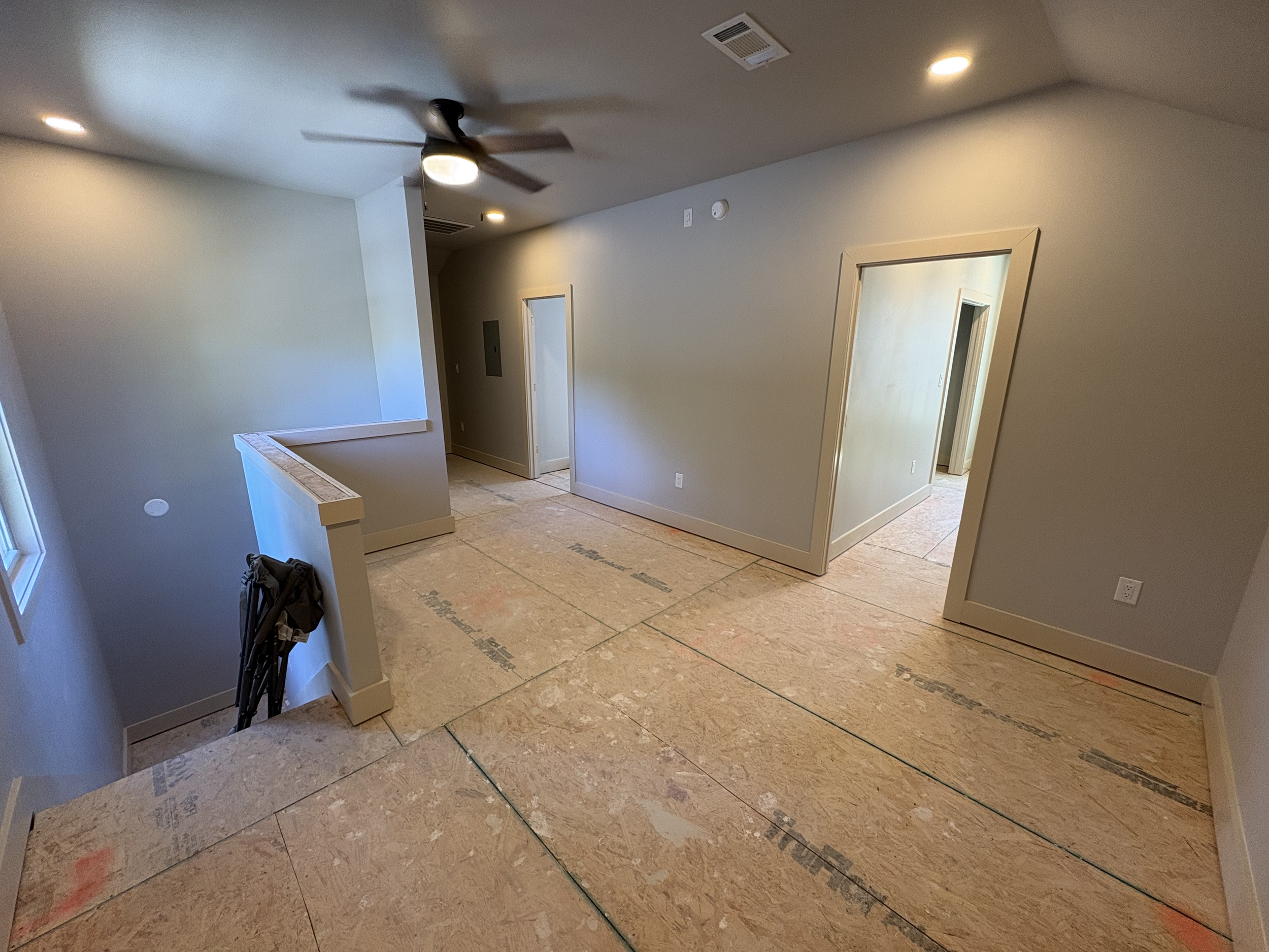 Interior of a house under construction or renovation, showing an unfinished floor with exposed plywood, light-colored walls, a ceiling fan, ceiling lights, and doorways leading to other rooms.