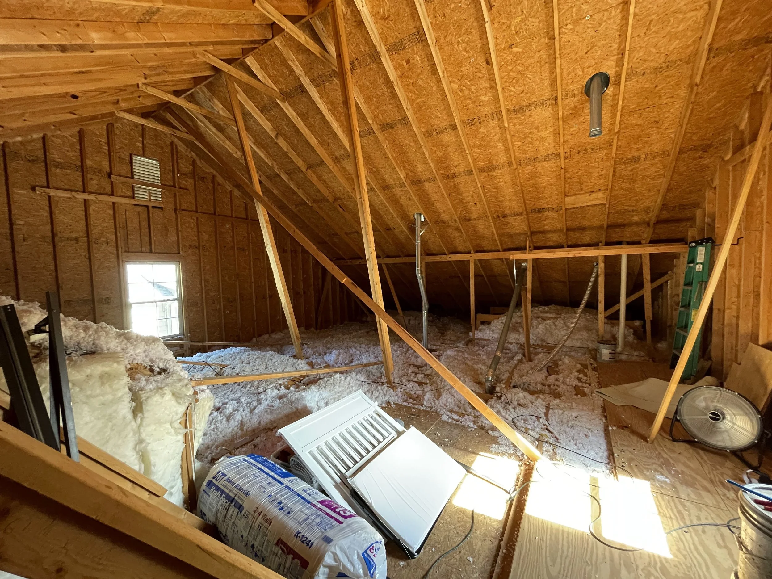 Attic under renovation with insulation, partially unfinished wooden framing, a small window, ladder, fan, and construction materials.