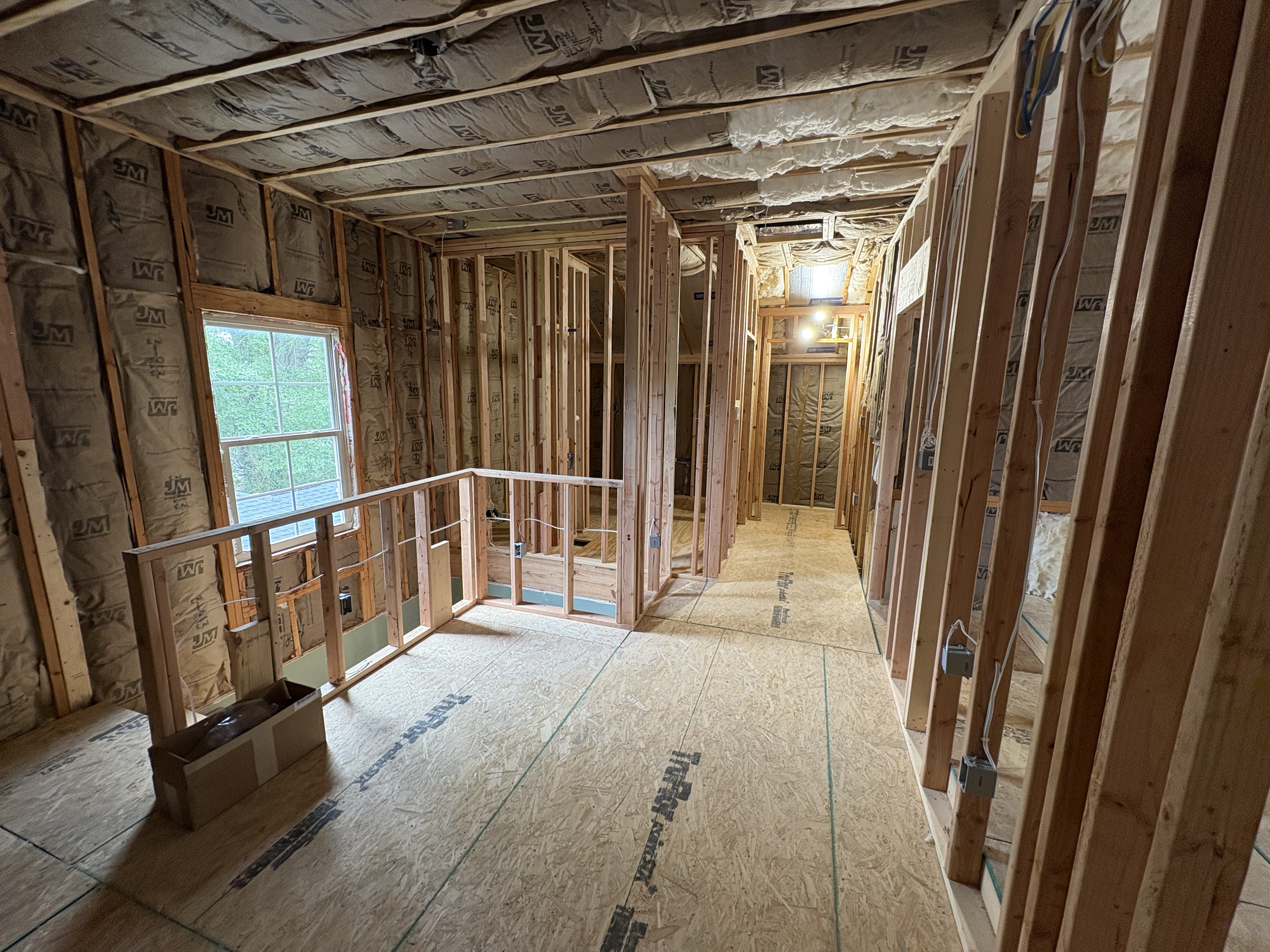 Interior view of a house under construction with exposed insulation, wooden framing, and plywood floors. A window on the left allows natural light in, and electrical wiring and outlets are visible along the studs.