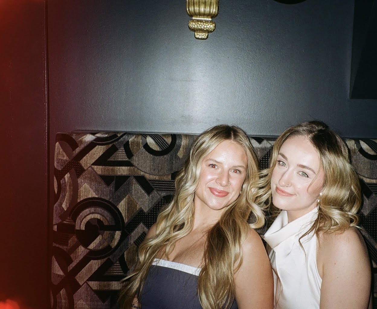 Two women with blonde hair smiling and sitting on a patterned booth seat in a dark-colored room, illuminated by a ceiling light overhead.