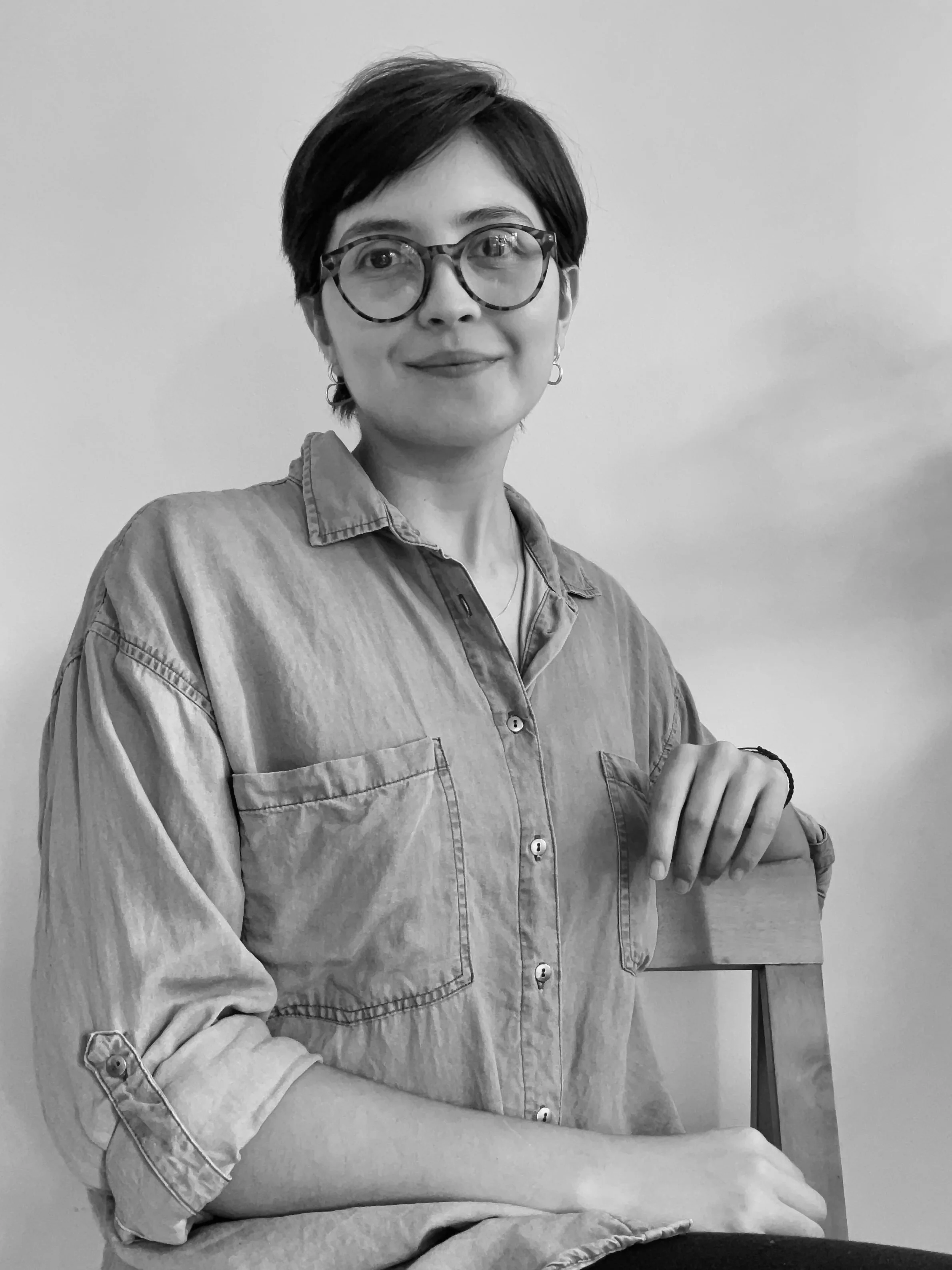 Retrato en blanco y negro de una mujer con gafas, cabello corto y oscuro, usando una camisa de mezclilla y sentada junto a una silla, con expresión tranquila y sonrisa suave.