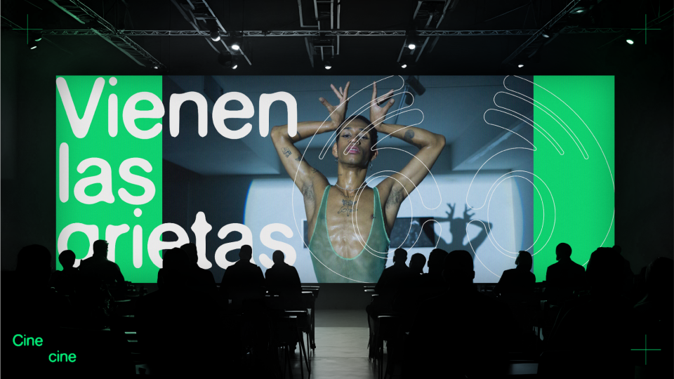 Proyección en una pantalla grande que dice "Vienen las ori*as" con una mujer en primer plano, sosteniéndose la cabeza, en un entorno de cine o evento, con público sentado en la oscuridad.