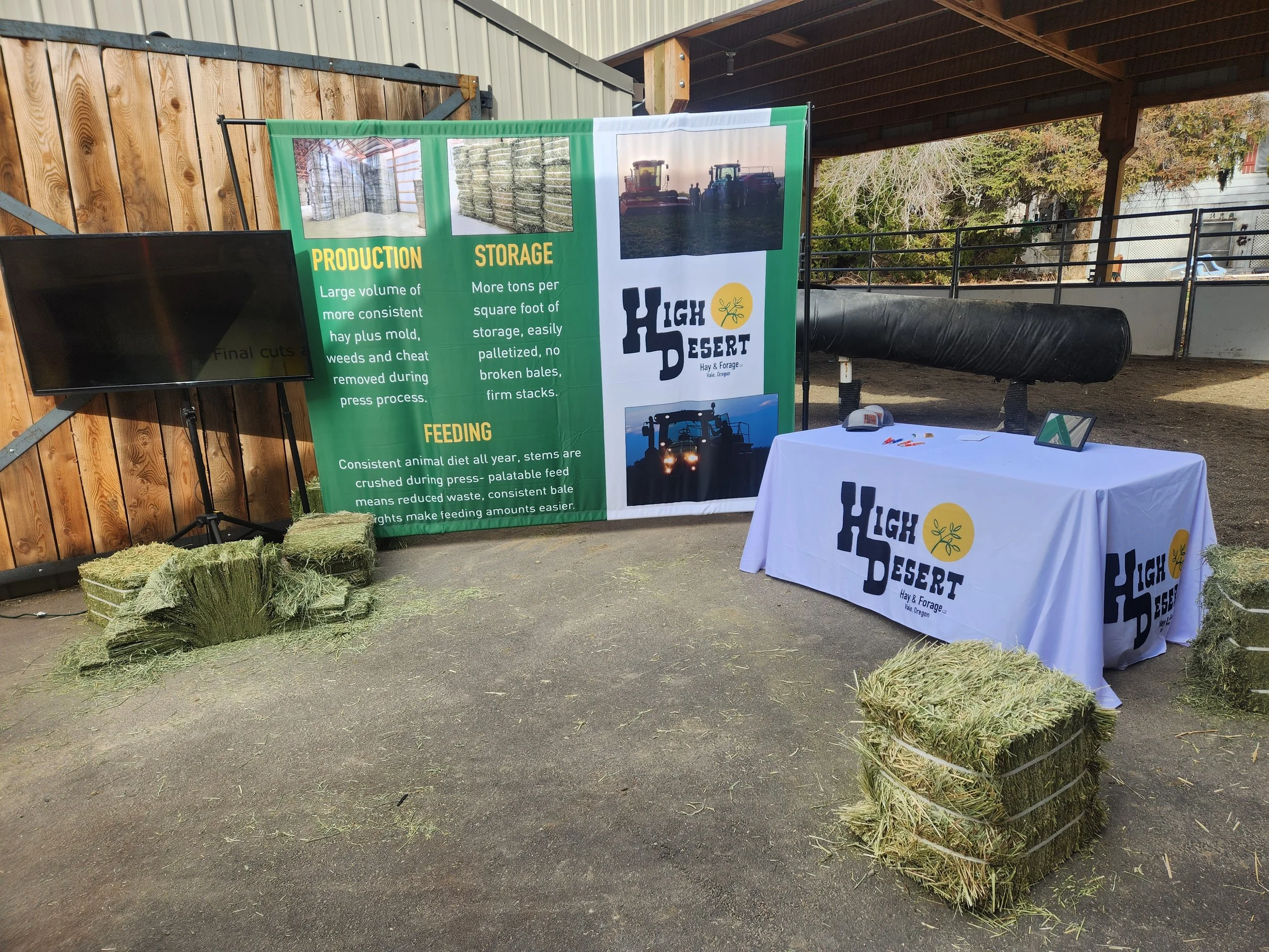 Exhibit booth at a farm and forage event with a green banner providing information about hay production, storage, and feeding. There are bales of hay displayed on the ground, a white table with the 'High Desert Hay & Forage' logo.
