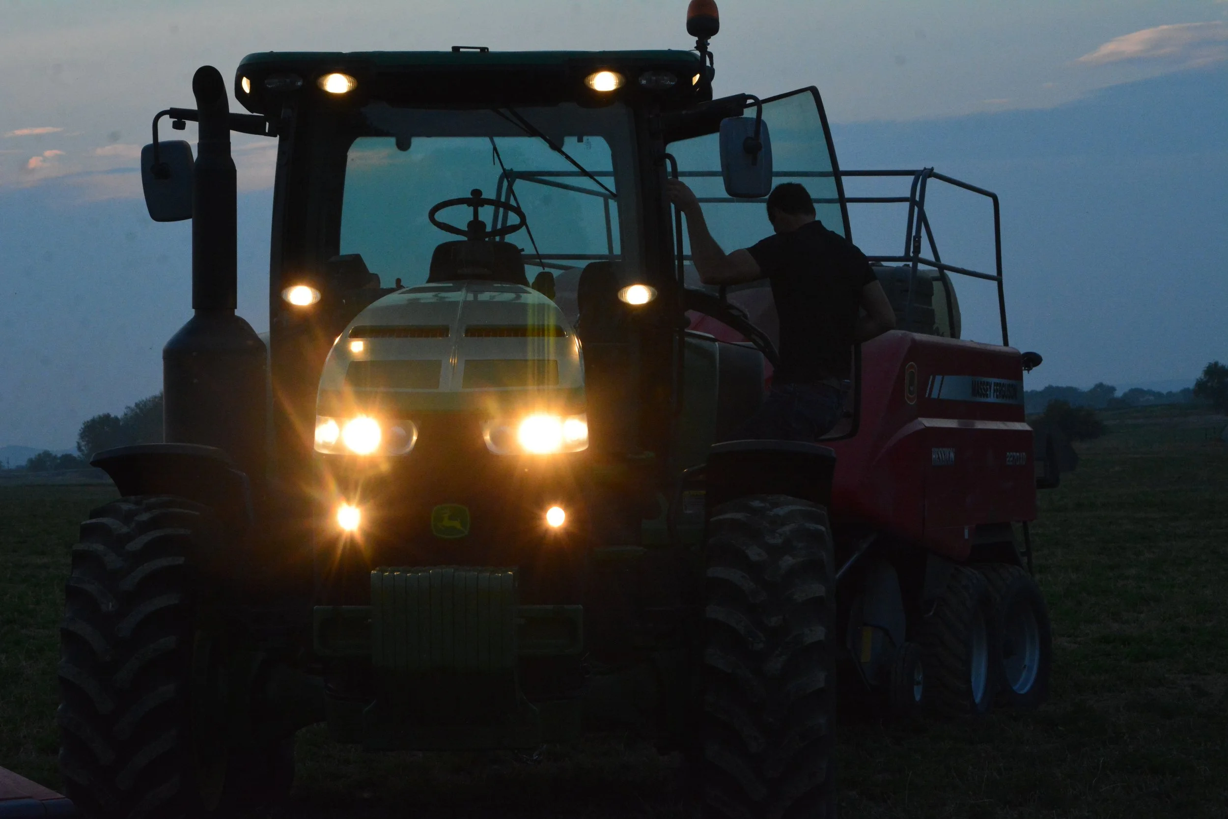 Farmer operating a tractor during dusk, with the tractor's headlights turned on, on an open field.