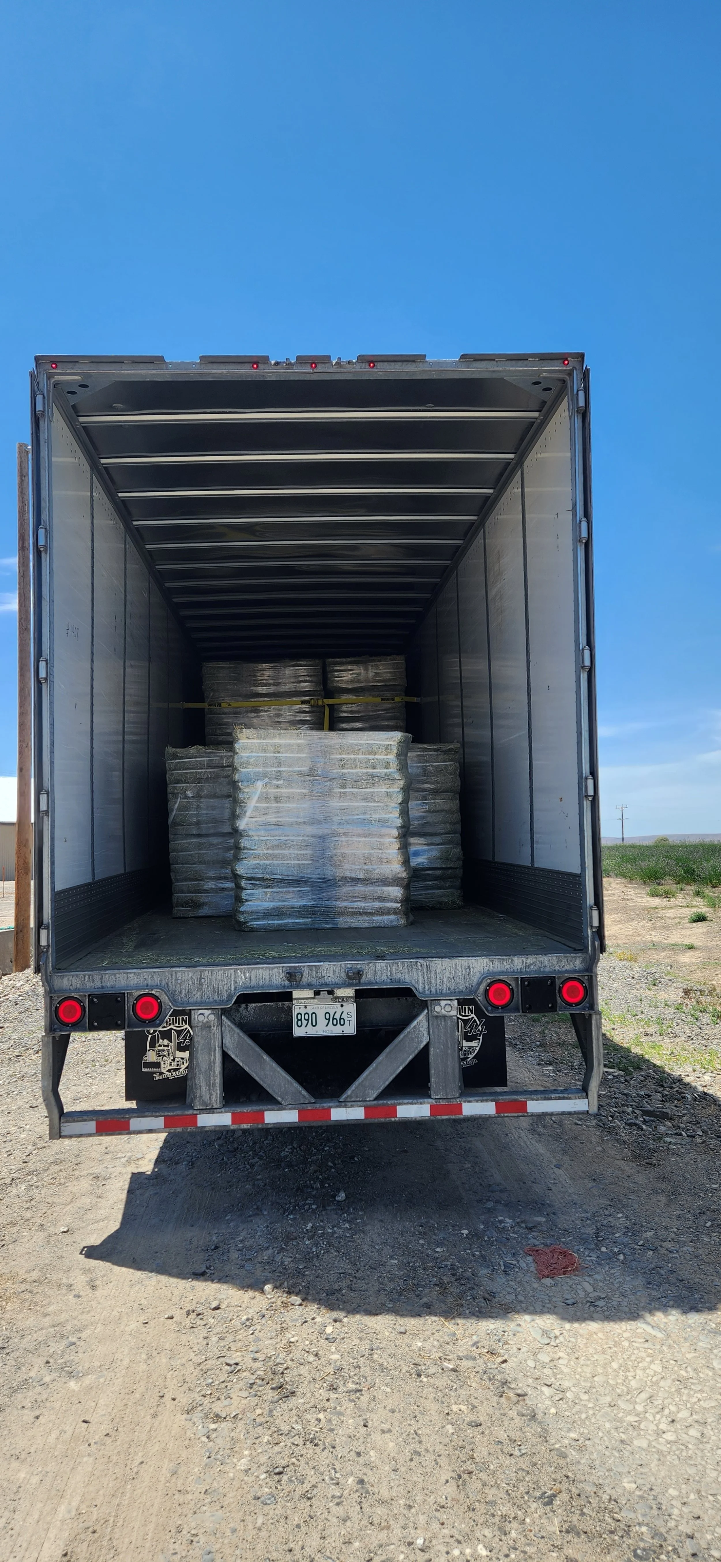 The back of a semi-truck trailer with pallets of stacked, wrapped material inside, parked on a dirt lot under a clear blue sky.