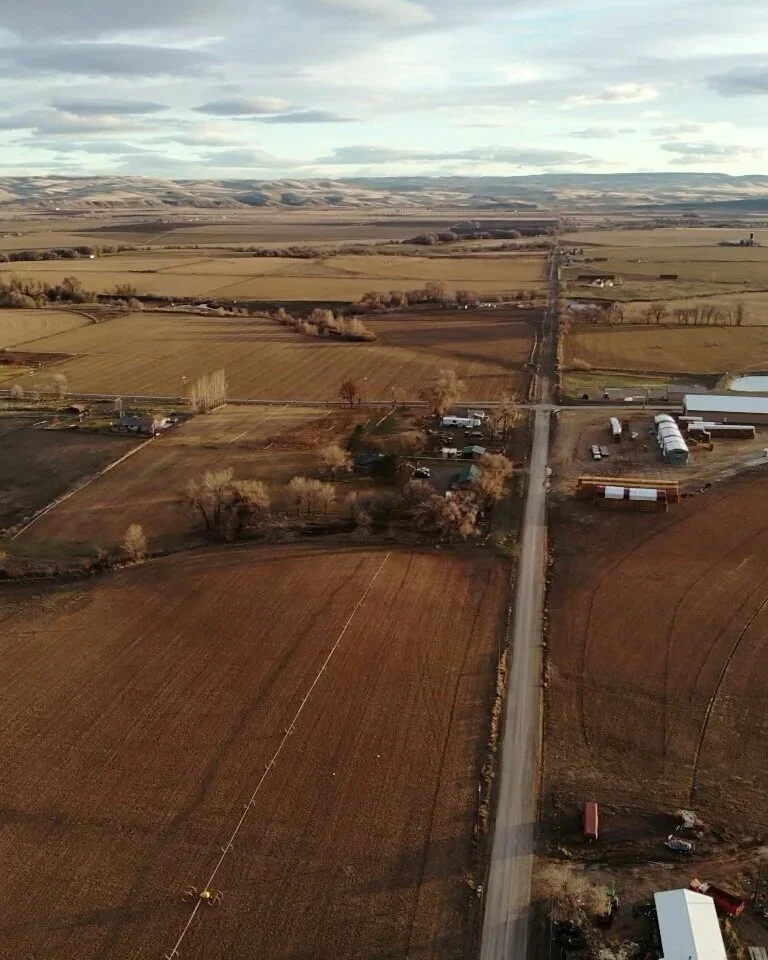 Aerial view of rural farmland with fields, farms, roads, and sparse trees under a partly cloudy sky.