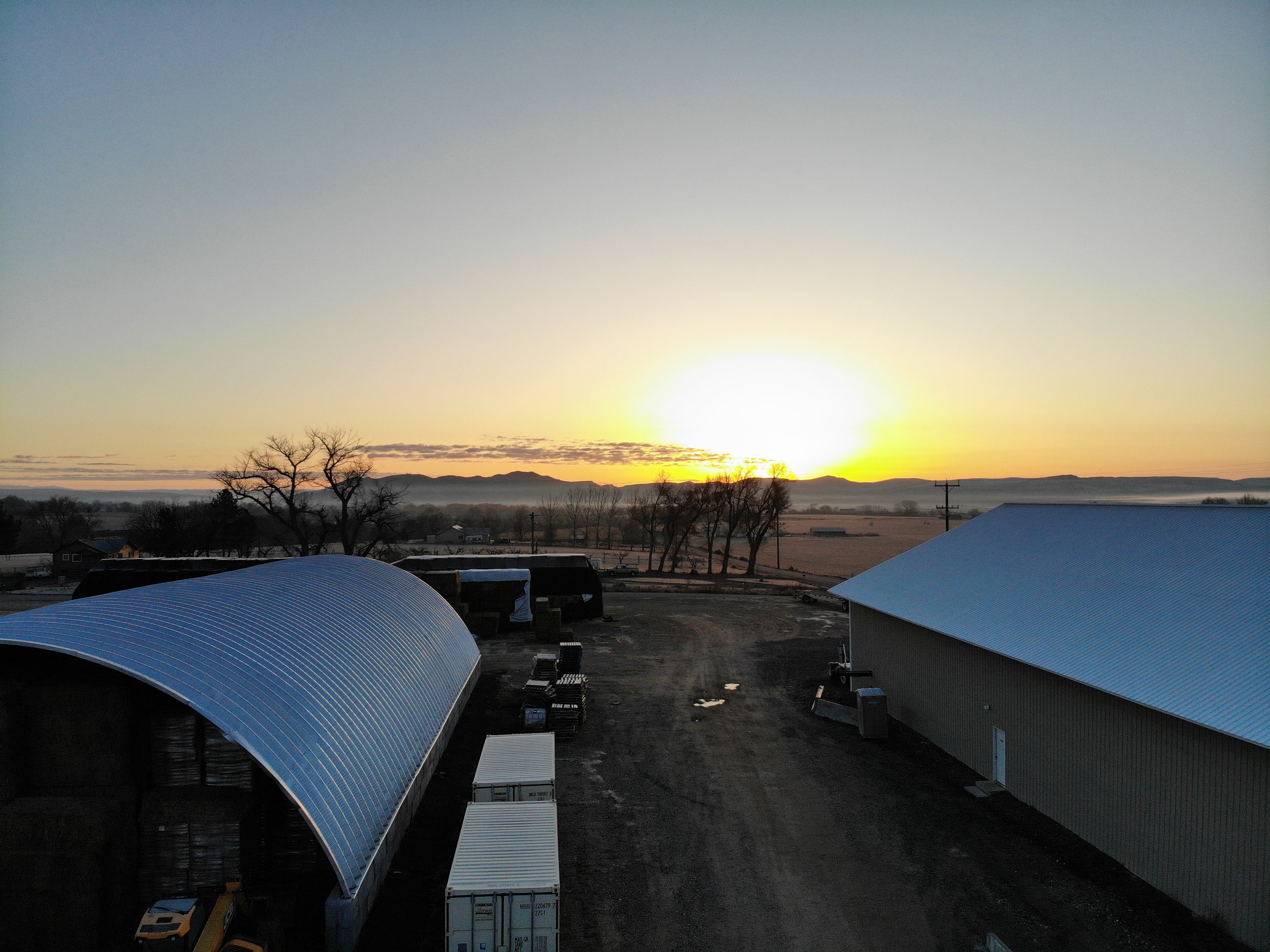 A rural farm scene during sunrise with large metal barns and trees, and distant mountains on the horizon.