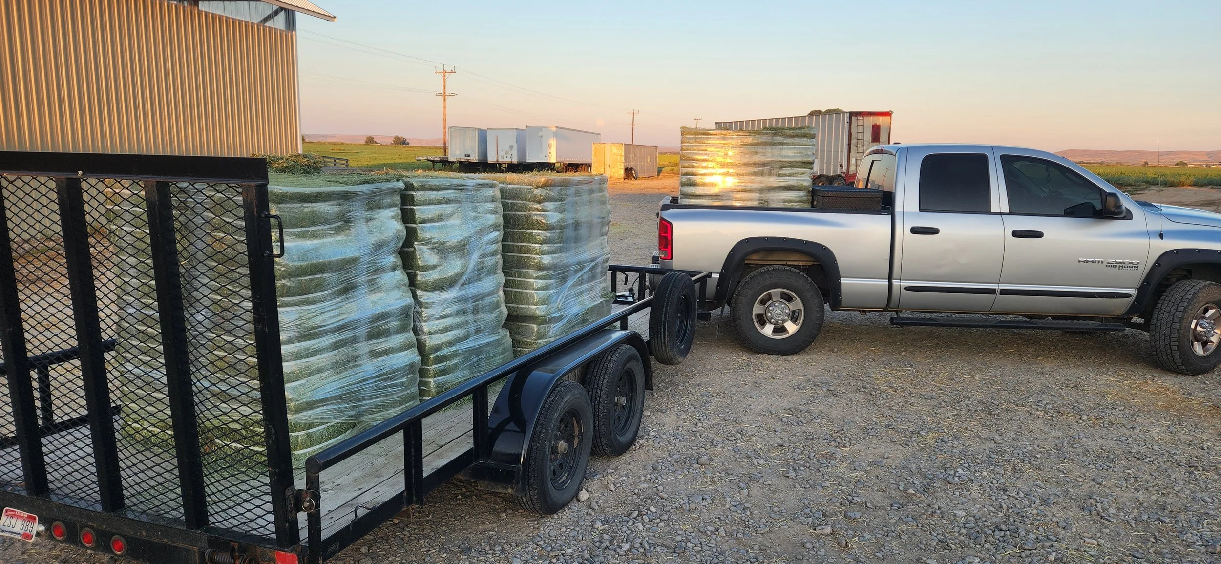 Silver pickup truck with black wheels parked next to a flatbed trailer loaded with large wrapped bales of hay, with a metal building to the left and open field in the background during sunset.