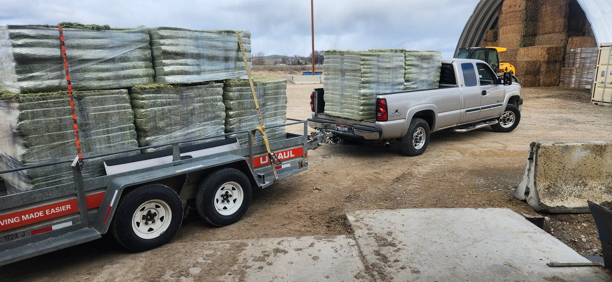 A silver pickup truck towing a small trailer filled with large wrapped hay bales, parked in a farmyard with a large barn filled with haystacks in the background.