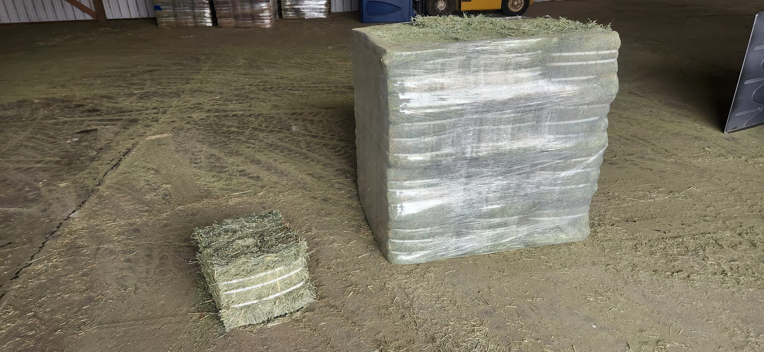 Large bundle and smaller bale of dried hay wrapped in clear plastic in a barn with concrete floor.