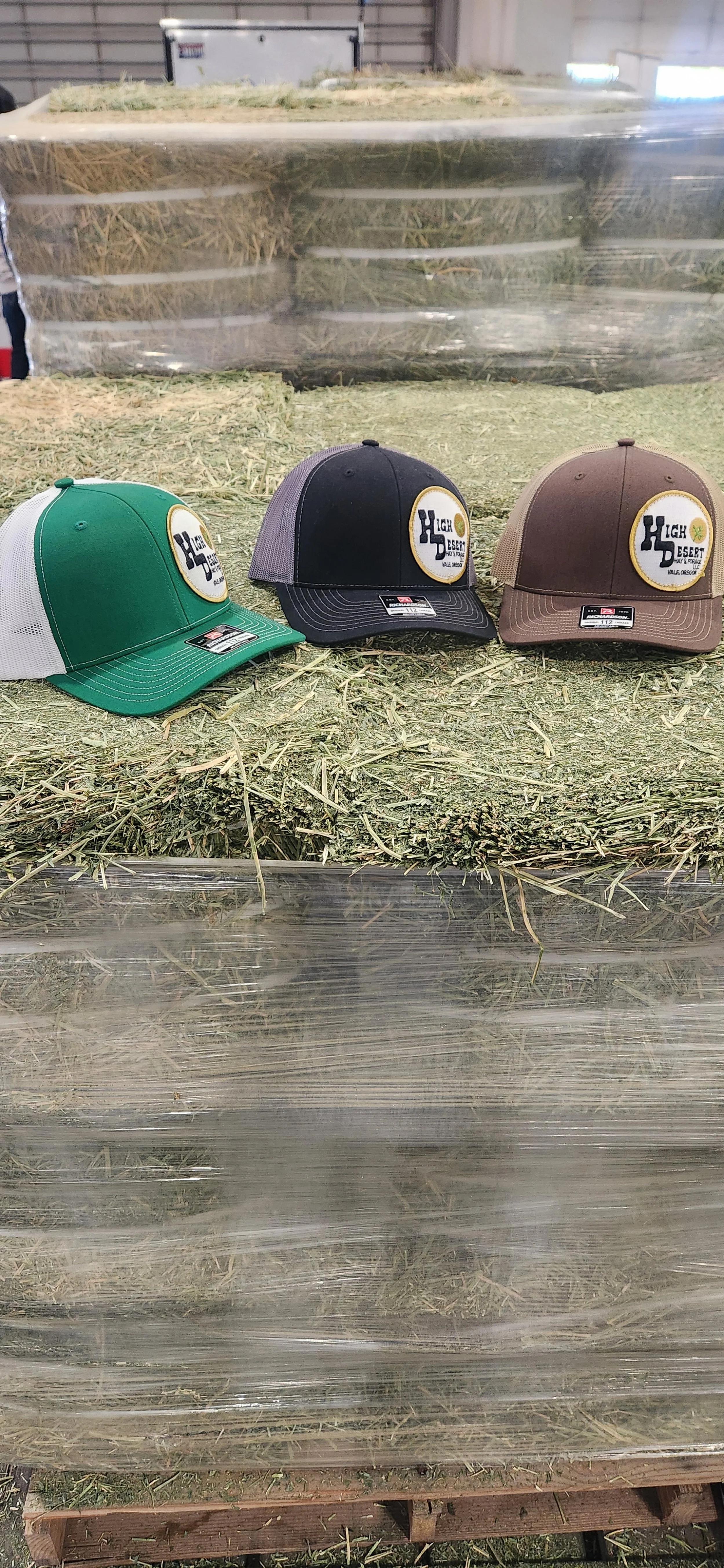 Three baseball caps with patches displaying the text 'High Desert Hay and Forage' on a bed of hay in a warehouse. The caps are in green with a white mesh back, black with a gray mesh back, and brown with a matching mesh back.