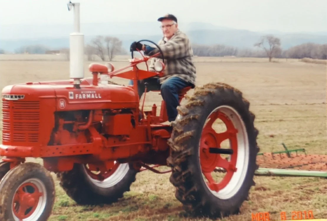 A man sitting on a red Farmall tractor in a field, with mountains and trees in the background.