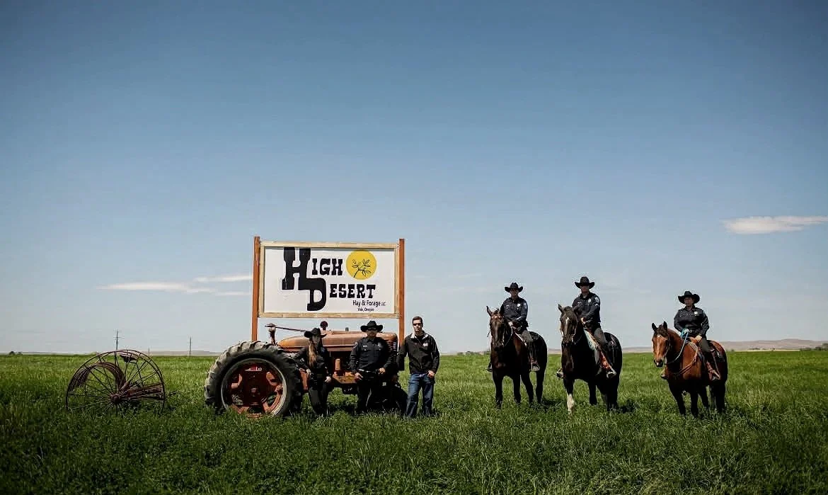 Group of five people, some on horseback and some standing, in a green field under a blue sky, with a sign reading High Desert Hay & Forage.