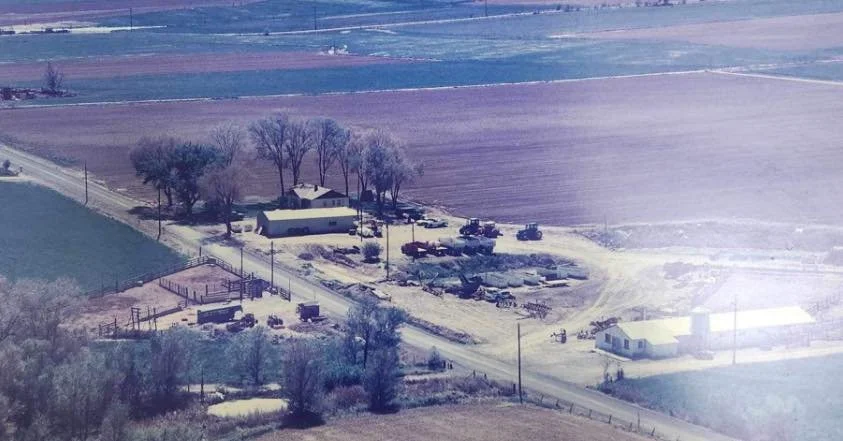 A rural farmstead with a house, barn, trees, and several farm vehicles and equipment, surrounded by fields.
