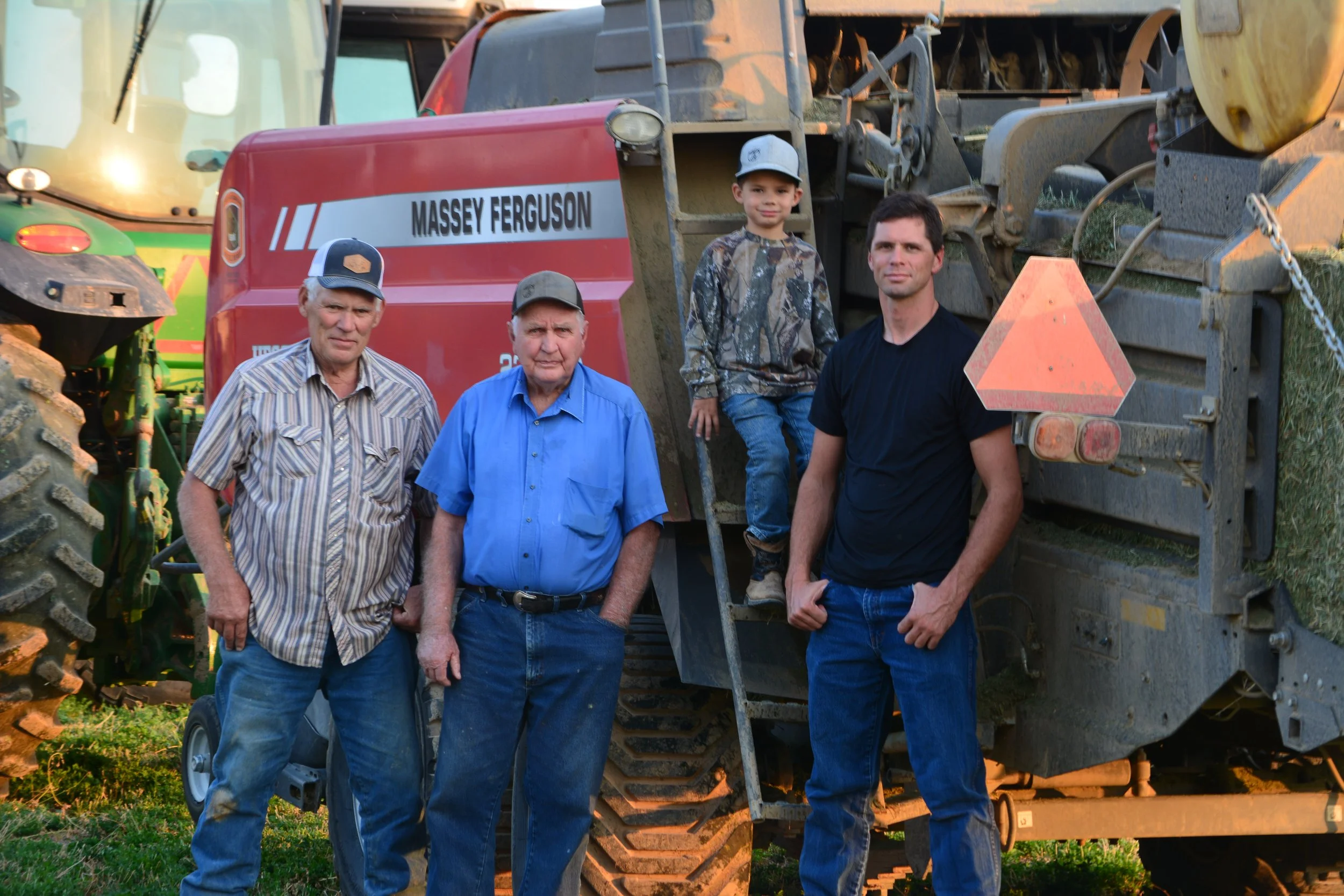 Four males, including a young boy, standing in front of a large Massey Ferguson baler in an outdoor rural setting during sunset.