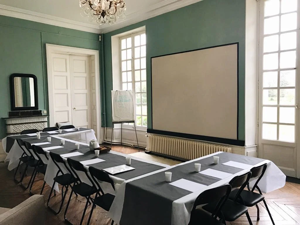 Salle de réunion avec table en U, chaises noires, papiers, tasses en papier, grand écran blanc, fenêtres avec lumière naturelle, miroir, et une cheminée.