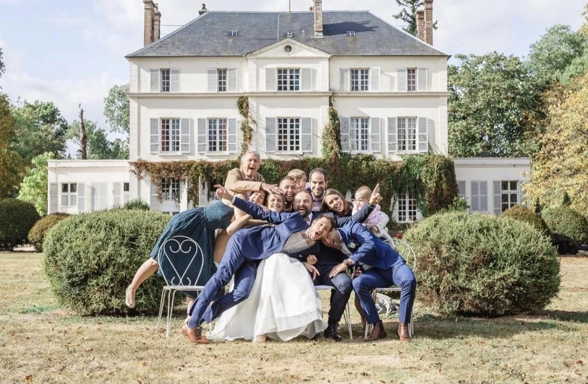 Groupe de personnes posant pour une photo de mariage devant une grande maison blanche avec jardins bien entretenus.