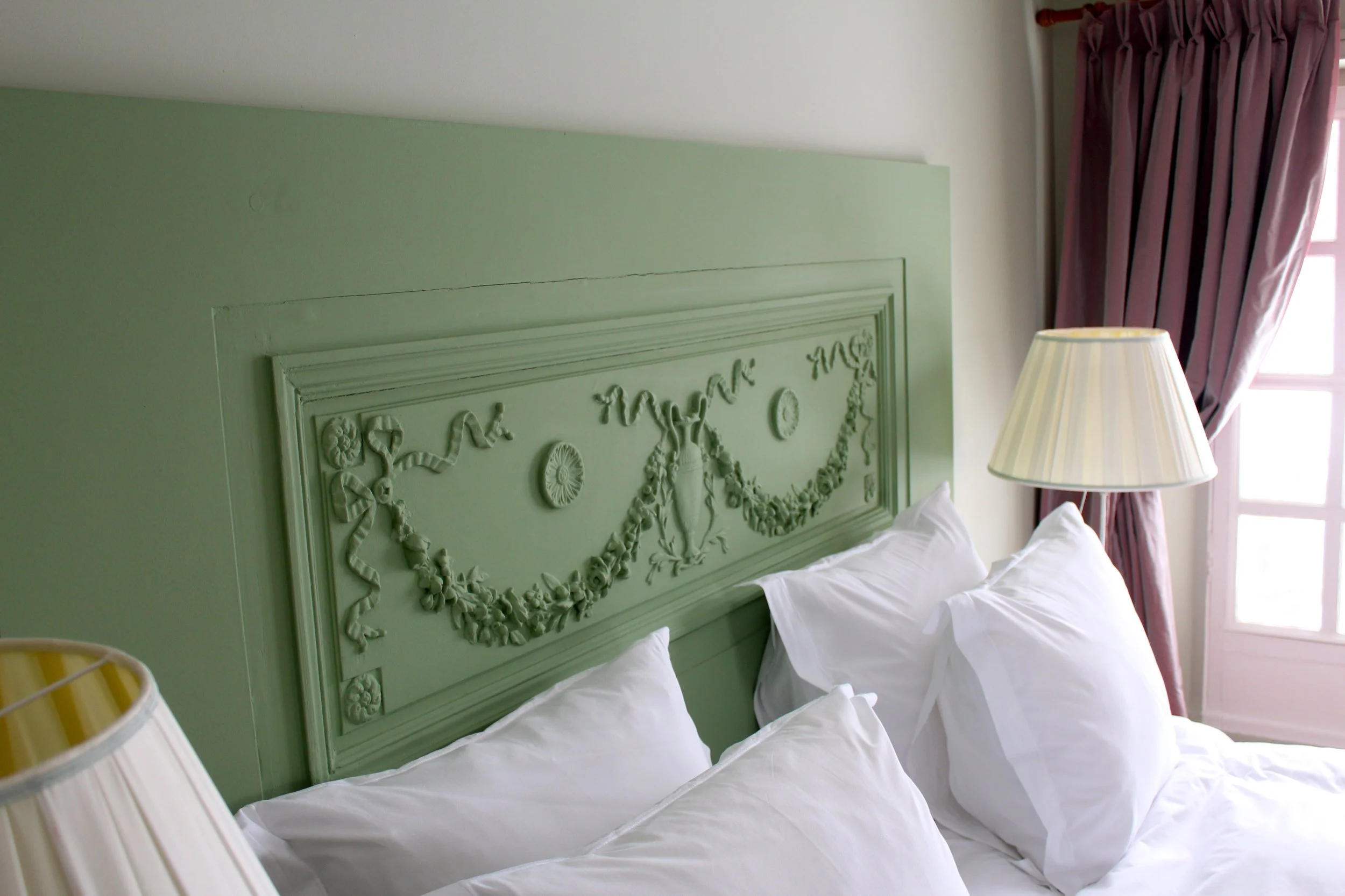 Lits avec de grands coussins blancs dans une chambre avec tête de lit verte ornée de motifs décoratifs et volets en bois pâle, éclairée par une lampe de chevet et une fenêtre avec rideaux violets.