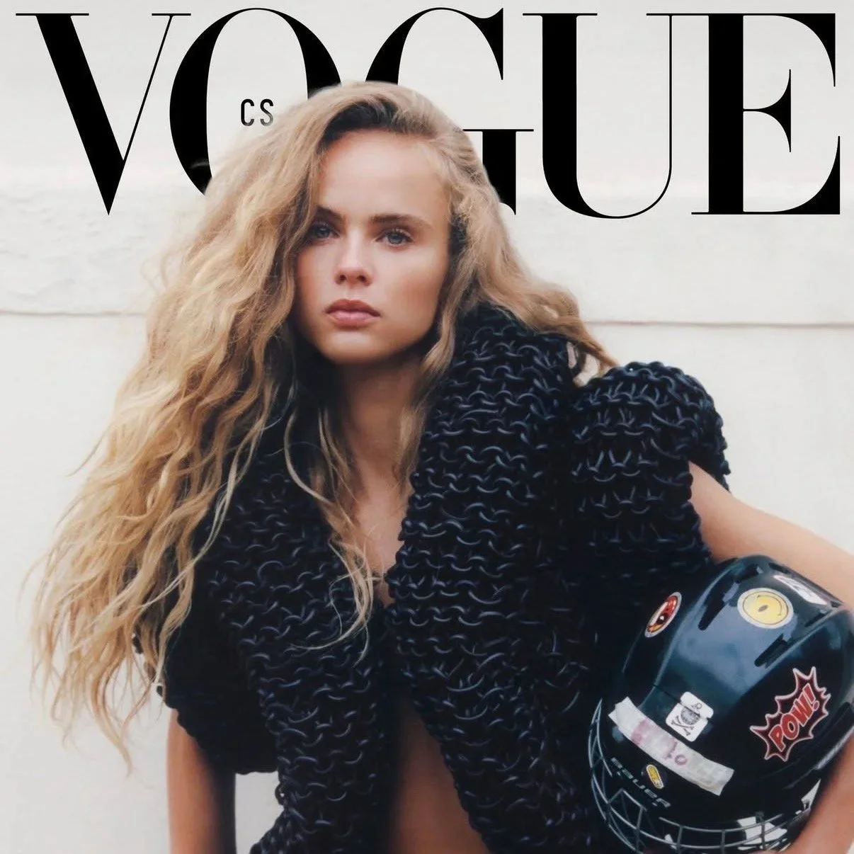 Une jeune femme aux cheveux longs et bouclés pose devant un mur blanc avec le logo Vogue. Elle porte un vêtement noir avec un casque de moto noir dans sa main.