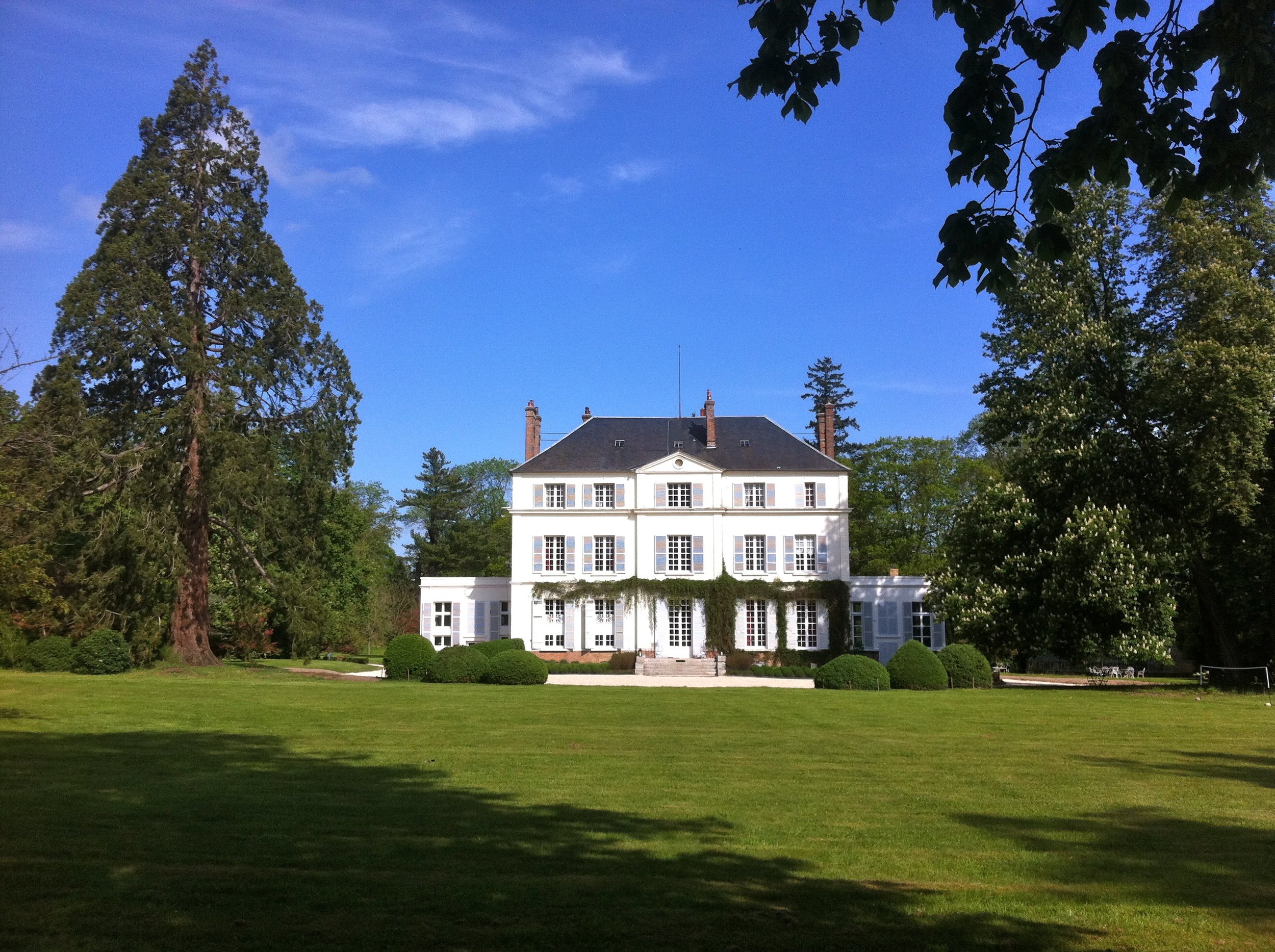 Un grand manoir blanc entouré d'arbres et d'un jardin verdoyant, sous un ciel bleu.