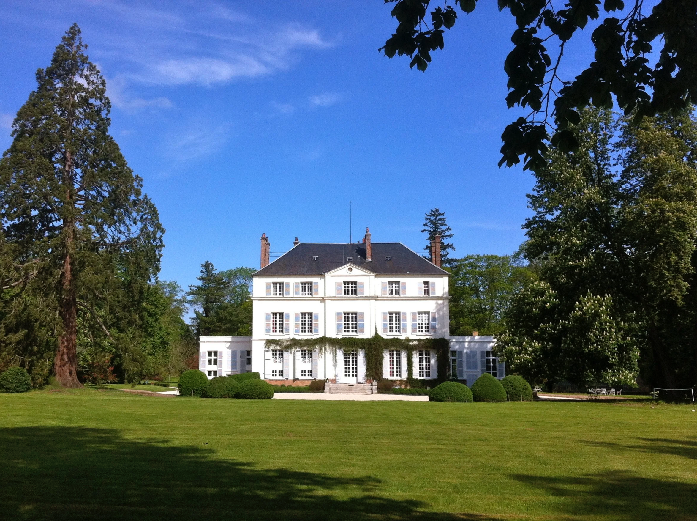 Un grand Château blanc avec plusieurs fenêtres, entouré d'arbres et de buissons verts, sous un ciel bleu clair.