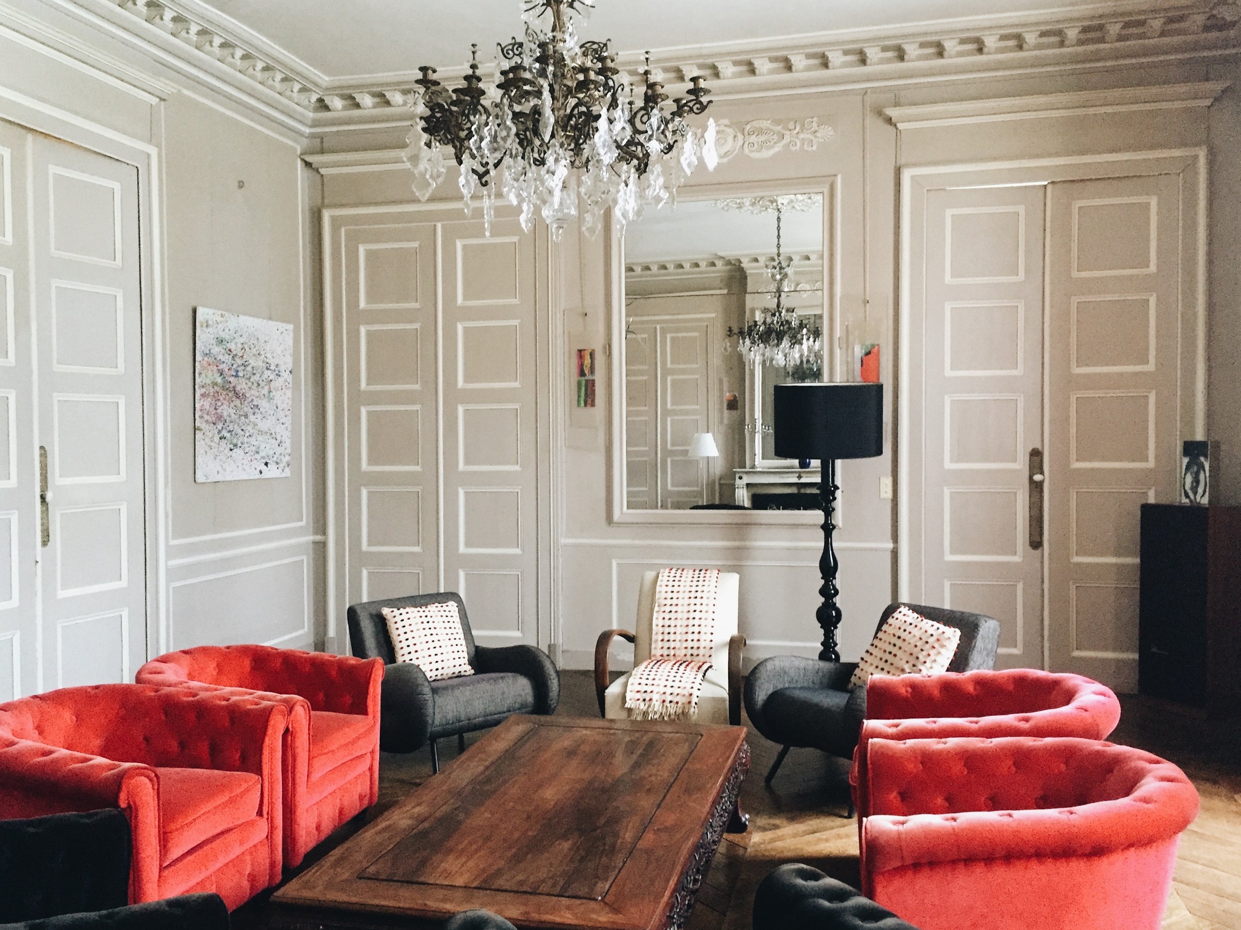 Salon élégant avec canapés rouges et gris autour d'une table en bois, grand miroir sur le mur, chandelier en cristal suspendu, lampadaire noir, murs ornés de moulures classiques.