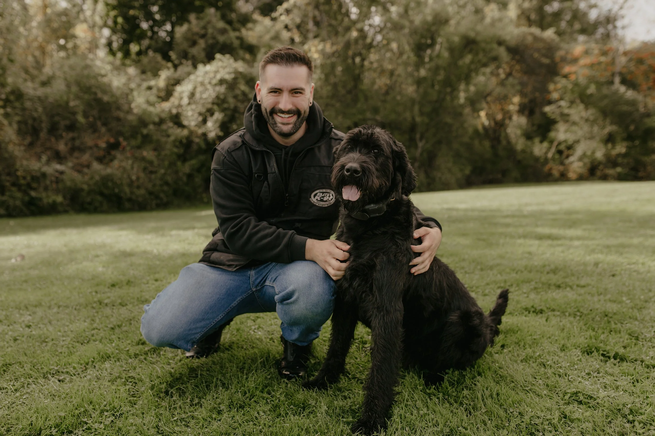 A man crouching on grass with a black dog in a park with trees in the background. The man is smiling, wearing a black jacket and blue jeans, holding the dog, which has its tongue out.