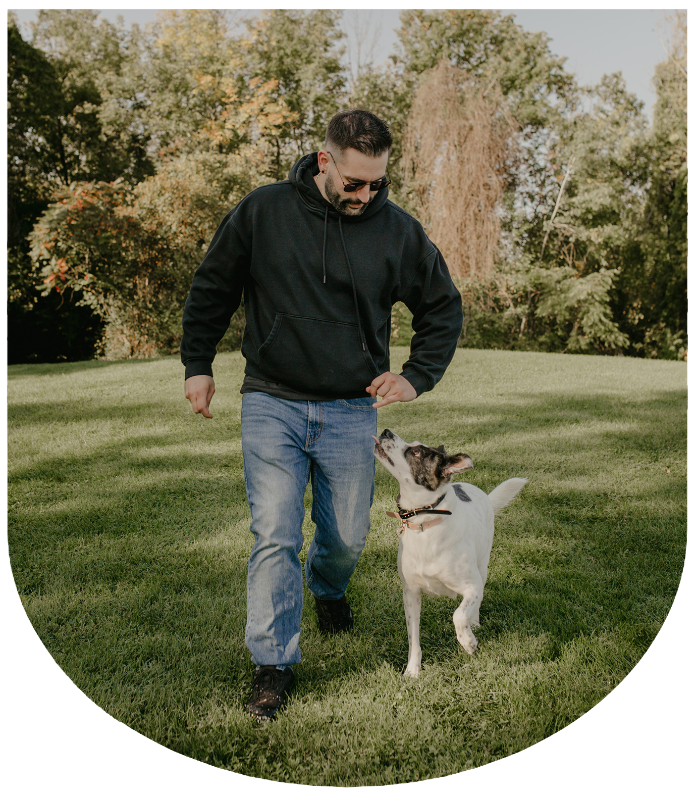 A man wearing sunglasses and a black hoodie playing with a dog in a park with green grass and trees in the background.