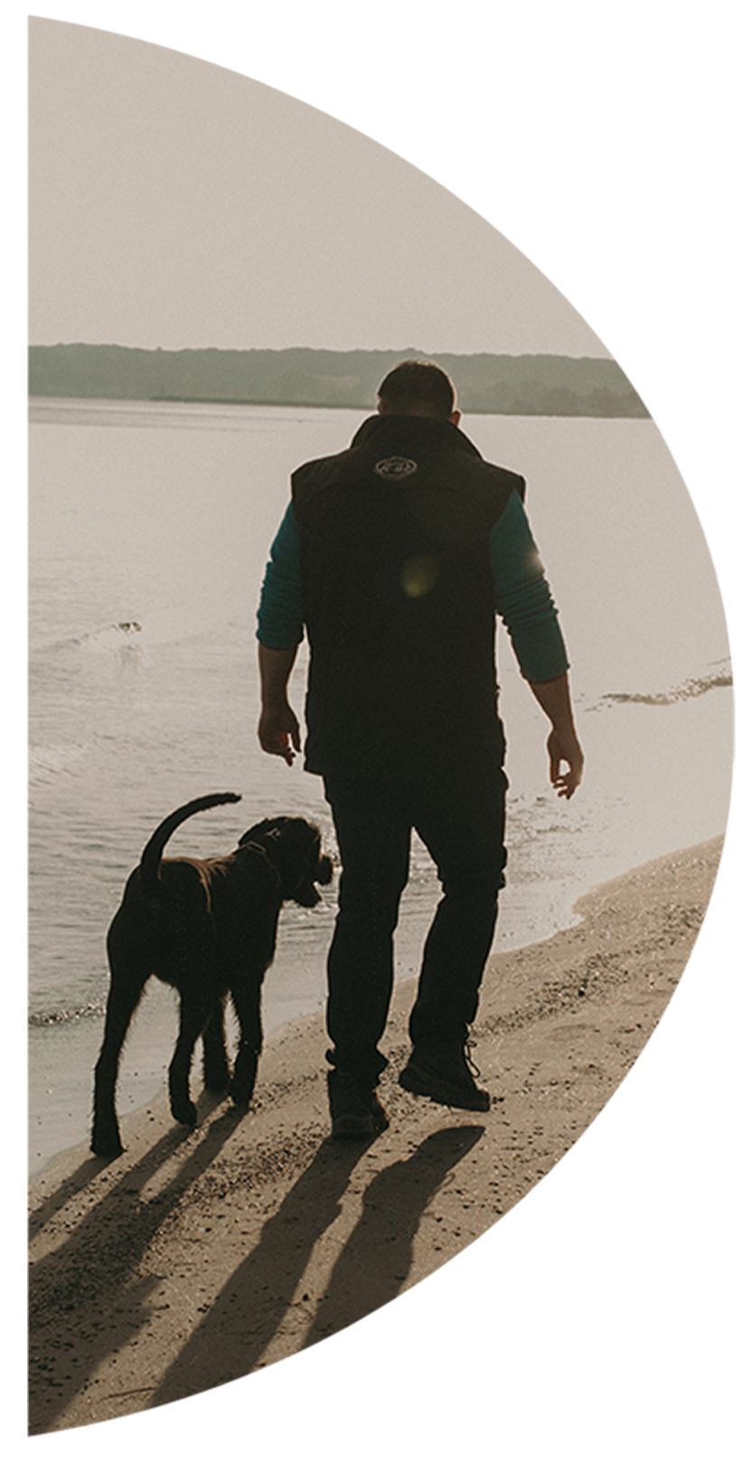A man walking along the beach with a dog near the water at sunset or sunrise.