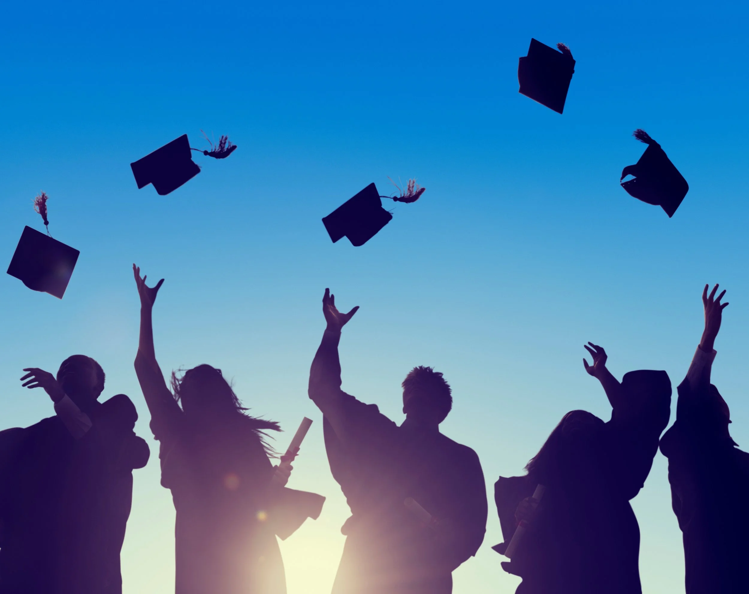 Silhouette of graduates tossing caps into the air against a bright blue sky.
