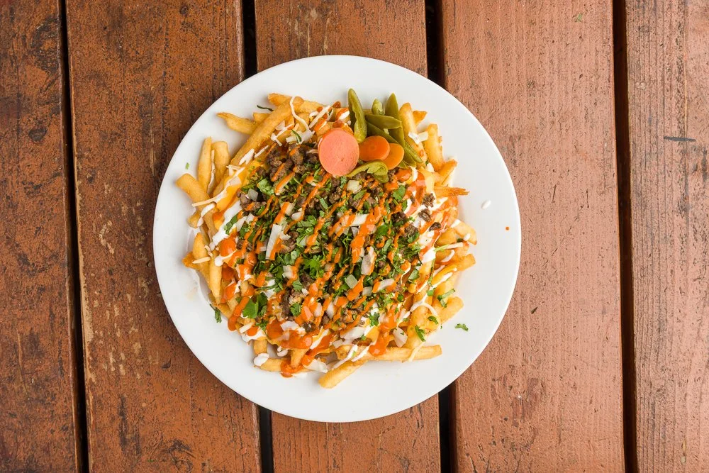 Plate of poutine with French fries, cheese curds, brown gravy, chopped green herbs, and pickles on a wooden table.