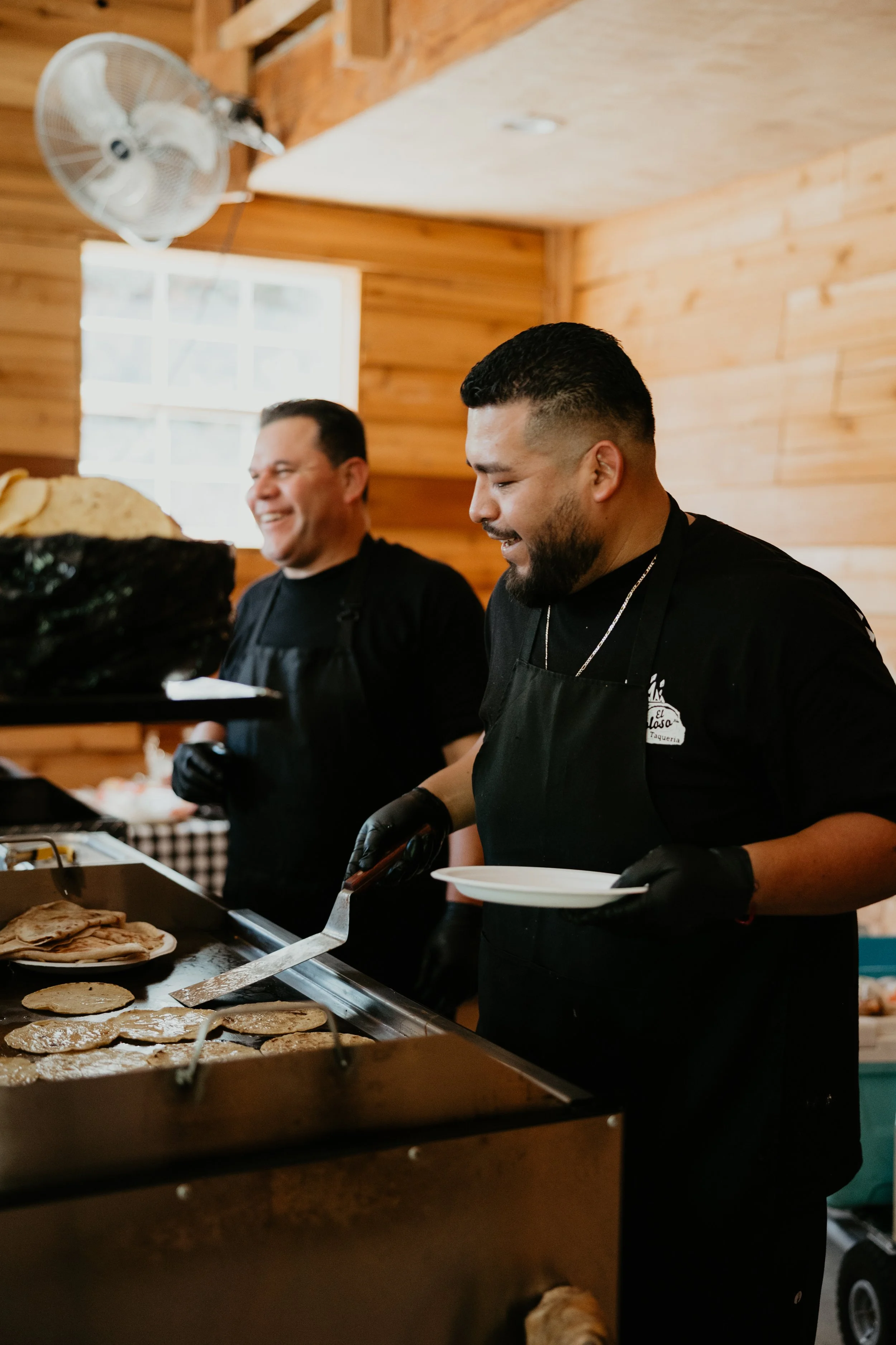 Two men cooking on a grill indoors; one man is flipping tortillas, the other holds a plate and smiling.