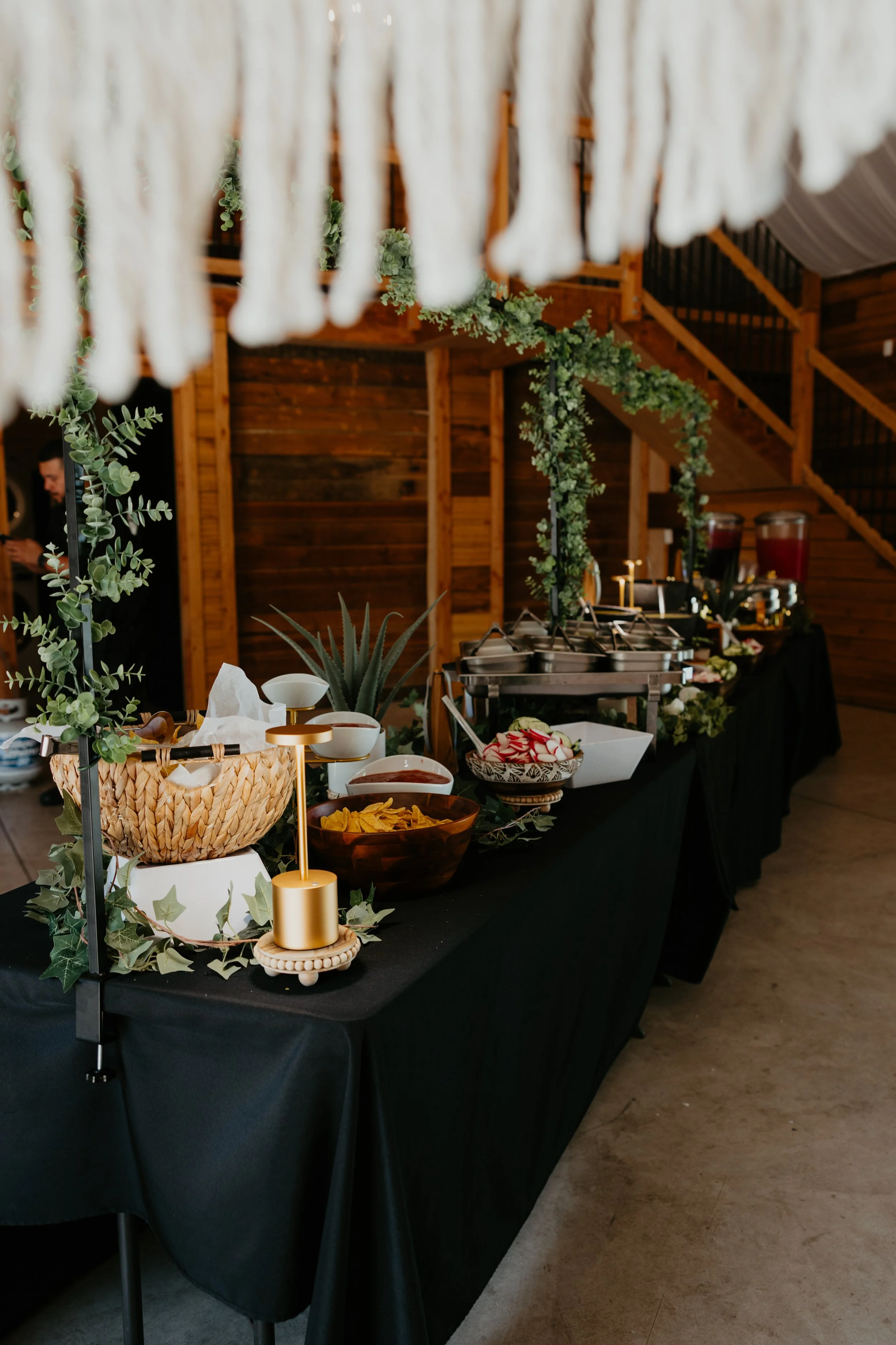 Buffet table with food and decorations inside a wooden venue, featuring a black tablecloth, greenery, and a cascading white fabric overhead.