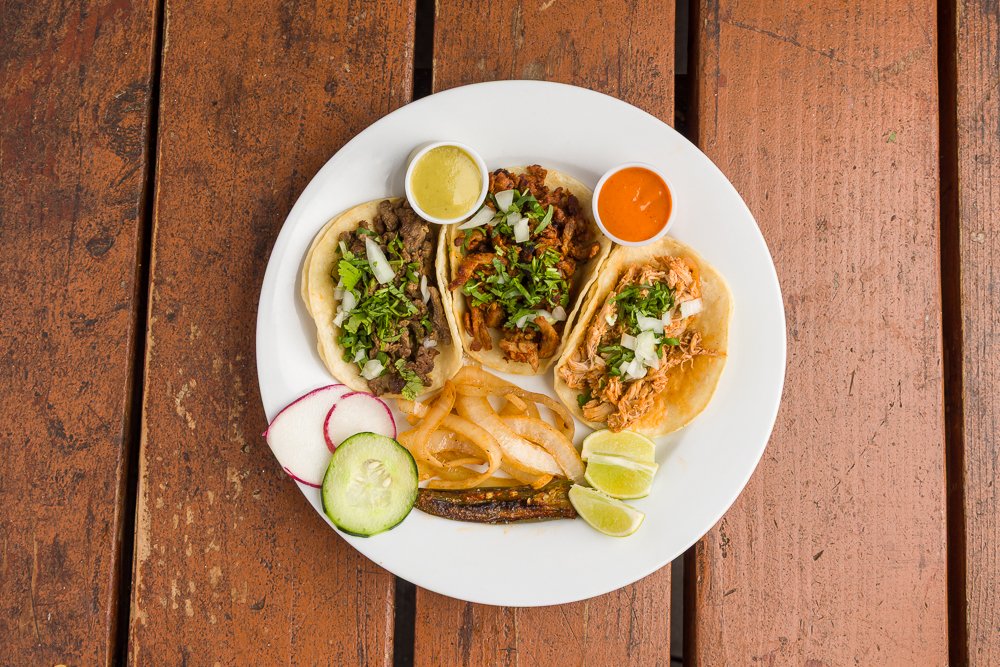 A white plate with three tacos topped with chopped onions and cilantro, garnished with lime wedges, sliced radishes, and onions. The plate also has salsa and green sauce, all on a wooden table.