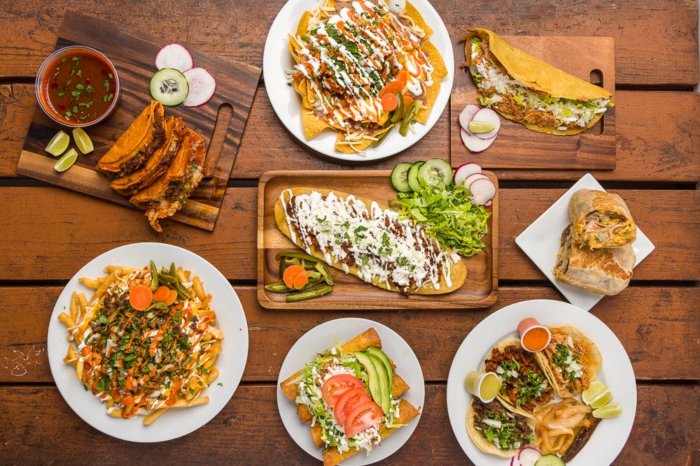A variety of Mexican dishes on a wooden table, including tacos, burritos, nachos, and a plate of Mexican street corn with lime wedges and radish slices.